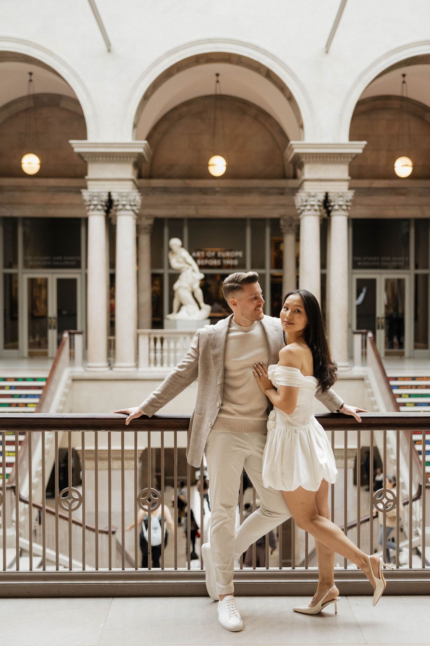 Stylish couple embracing on a museum balcony surrounded by neoclassical columns and a marble statue.