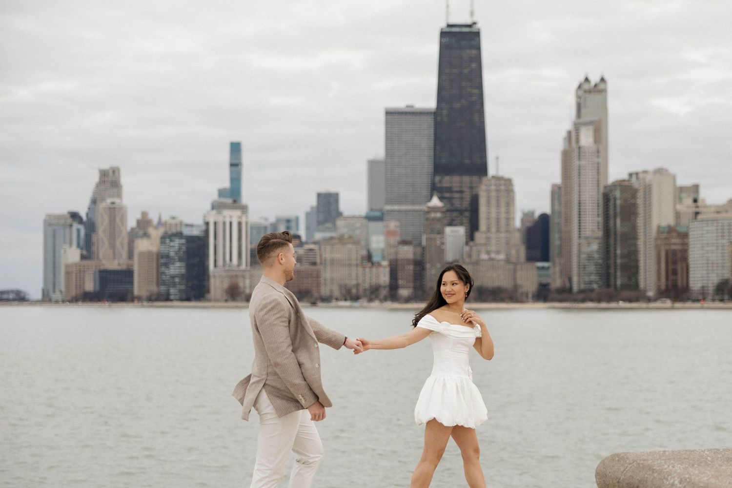 Couple holding hands walking near Lake Michigan with Chicago city skyline in the background.