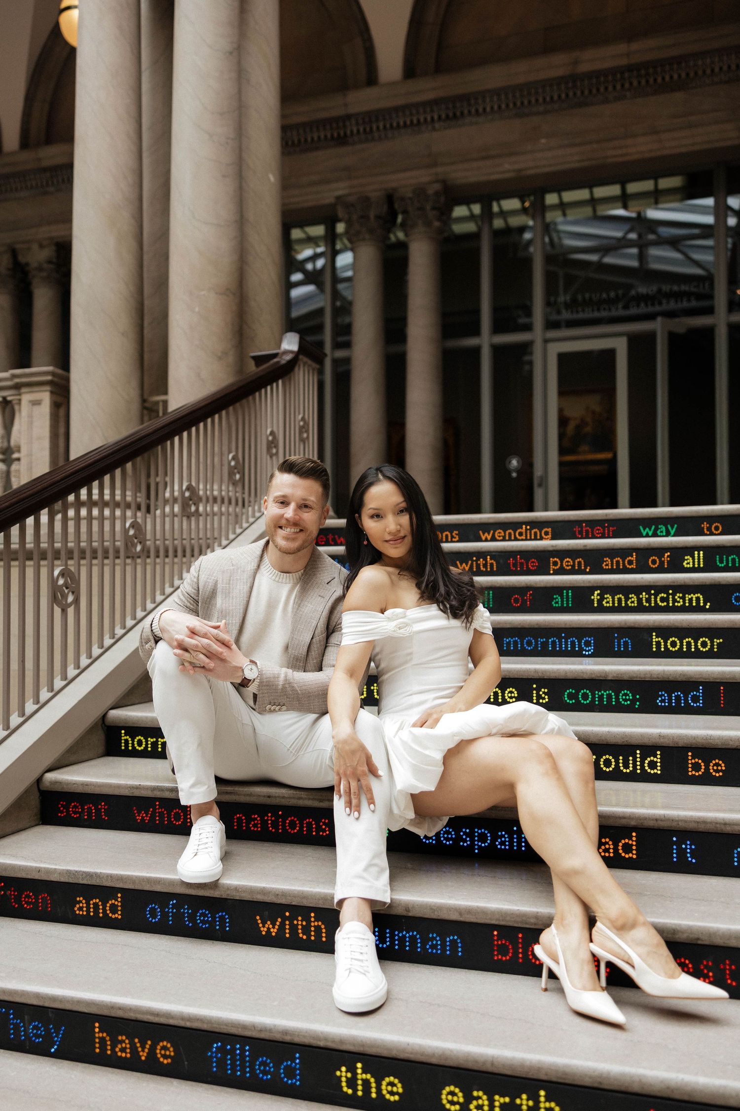 Couple sitting on decorative text-inscribed steps of elegant building with columns, man in beige suit, woman in white dress.