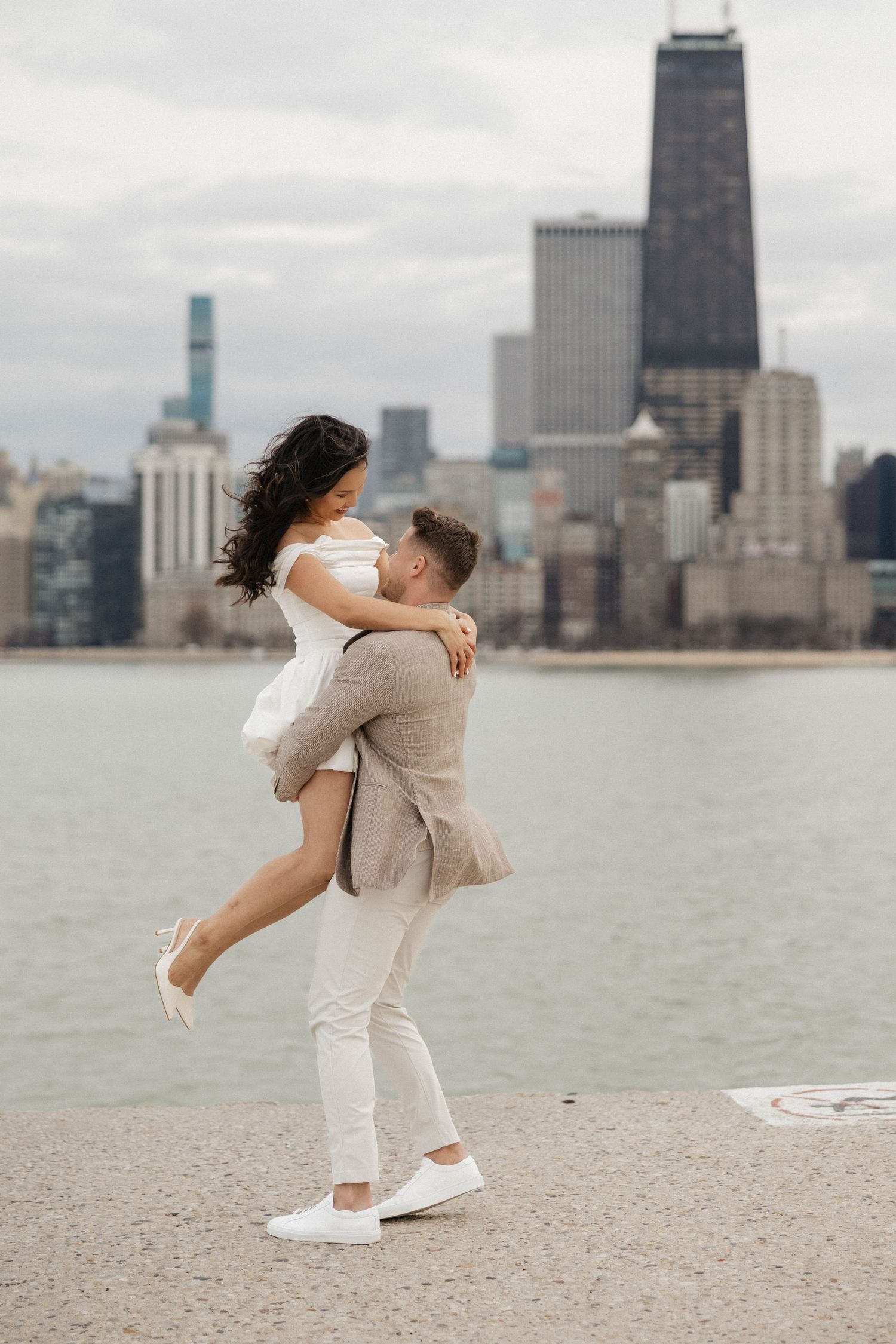 A couple embraces romantically on a lakefront with the Chicago skyline in the background on a cloudy day.