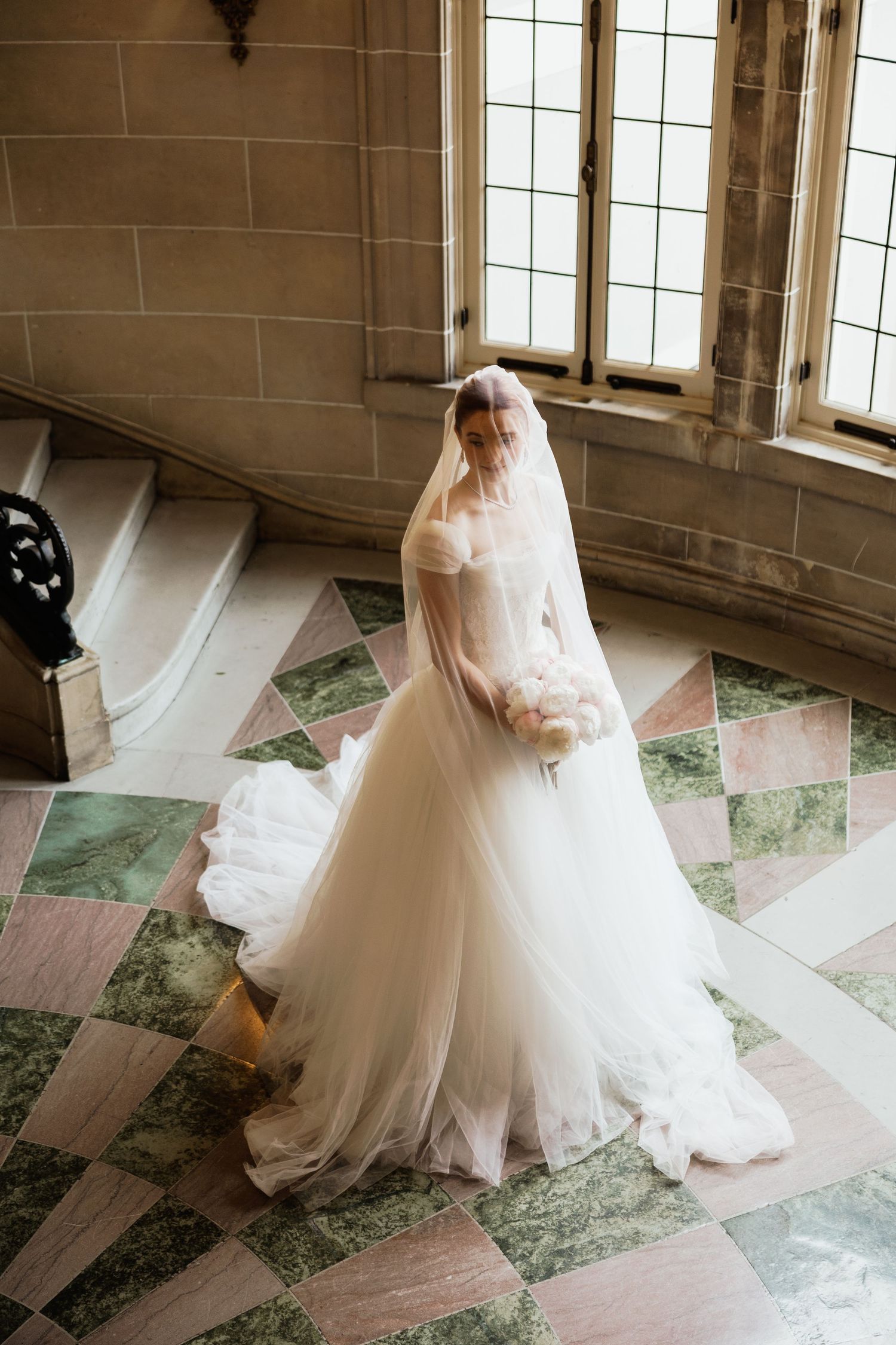 Bride in a flowing white tulle ball gown and long veil stands on marble checkered floor in an elegant stone building.