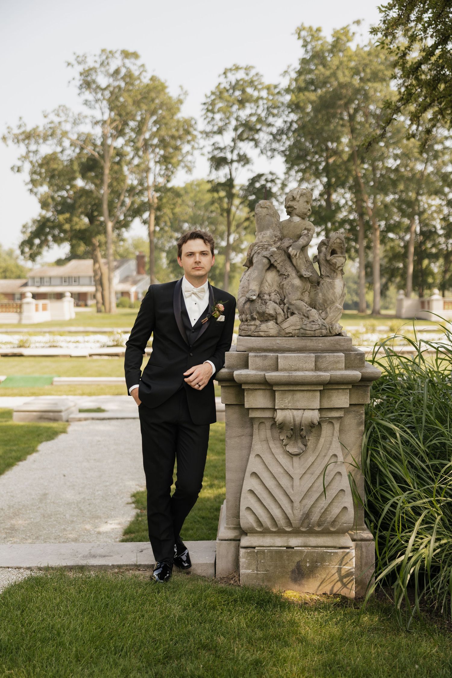 Groom in black tuxedo leans against ornate stone statue sculpture in lush garden.