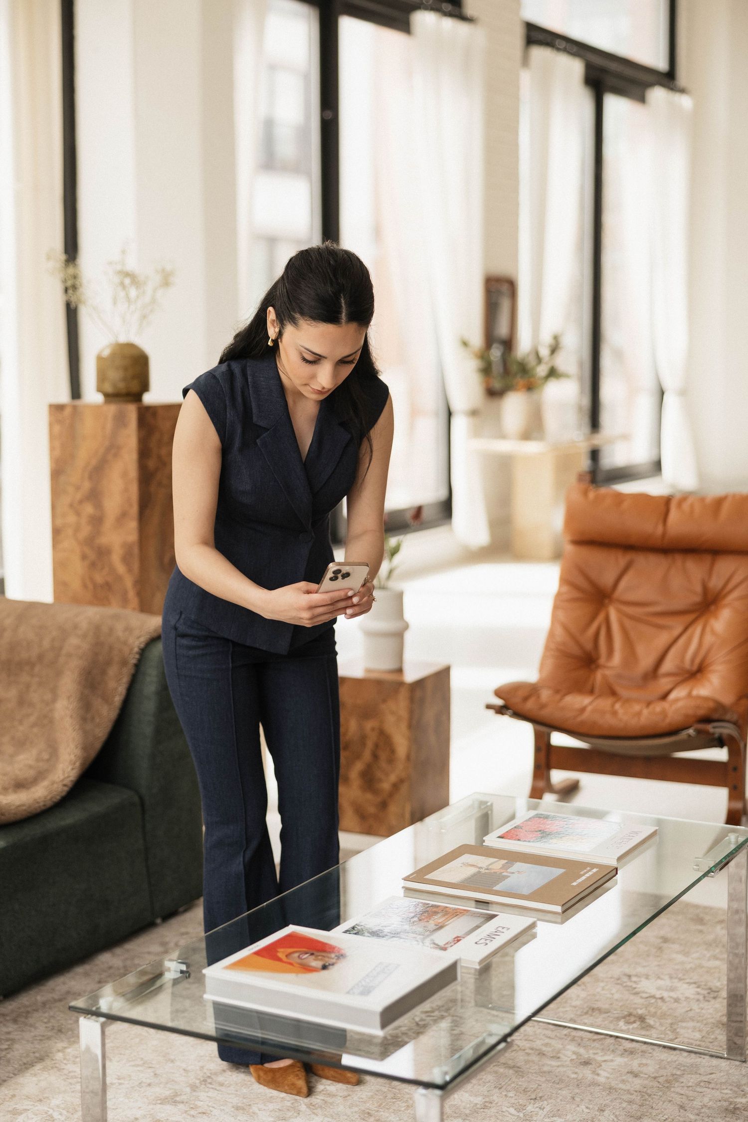 Woman in navy outfit photographs books on glass coffee table in modern living room with leather chair and large windows.