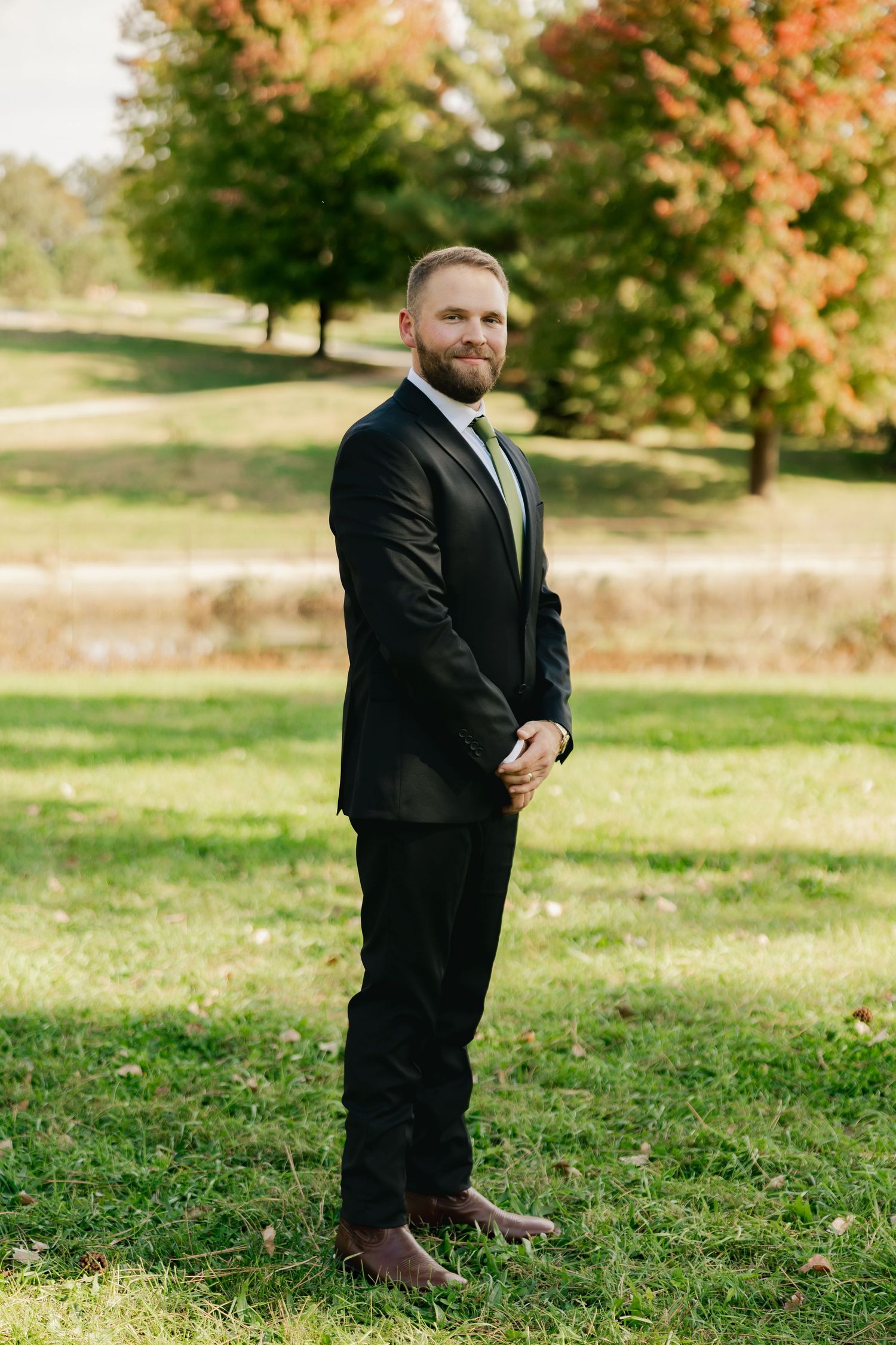 Bearded groom in a black suit, white shirt and green tie stands on a sunny grassy lawn with colorful autumn trees behind him.