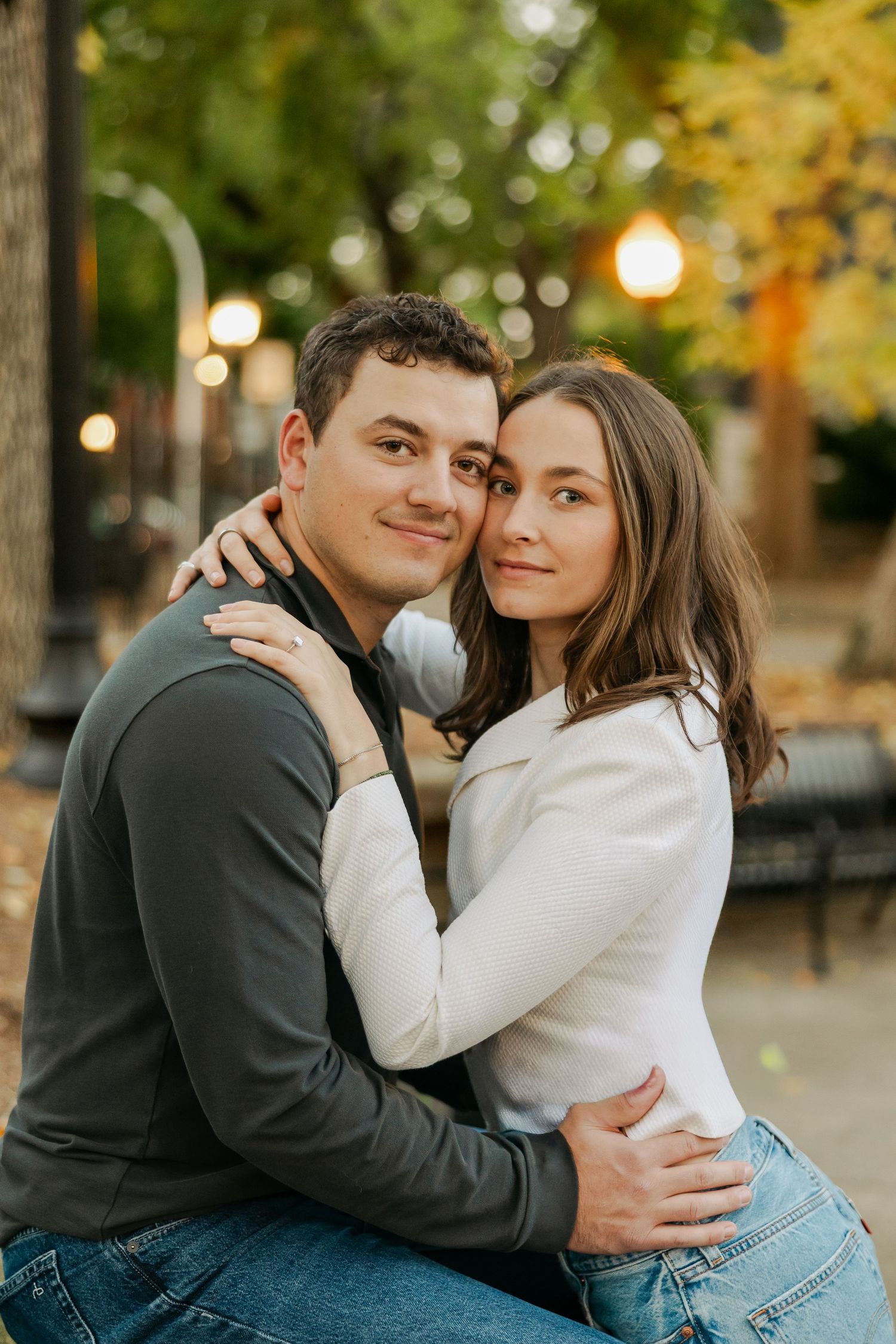 Smiling couple embracing closely on a park bench surrounded by autumn trees and glowing string lights during an engagement photo session.