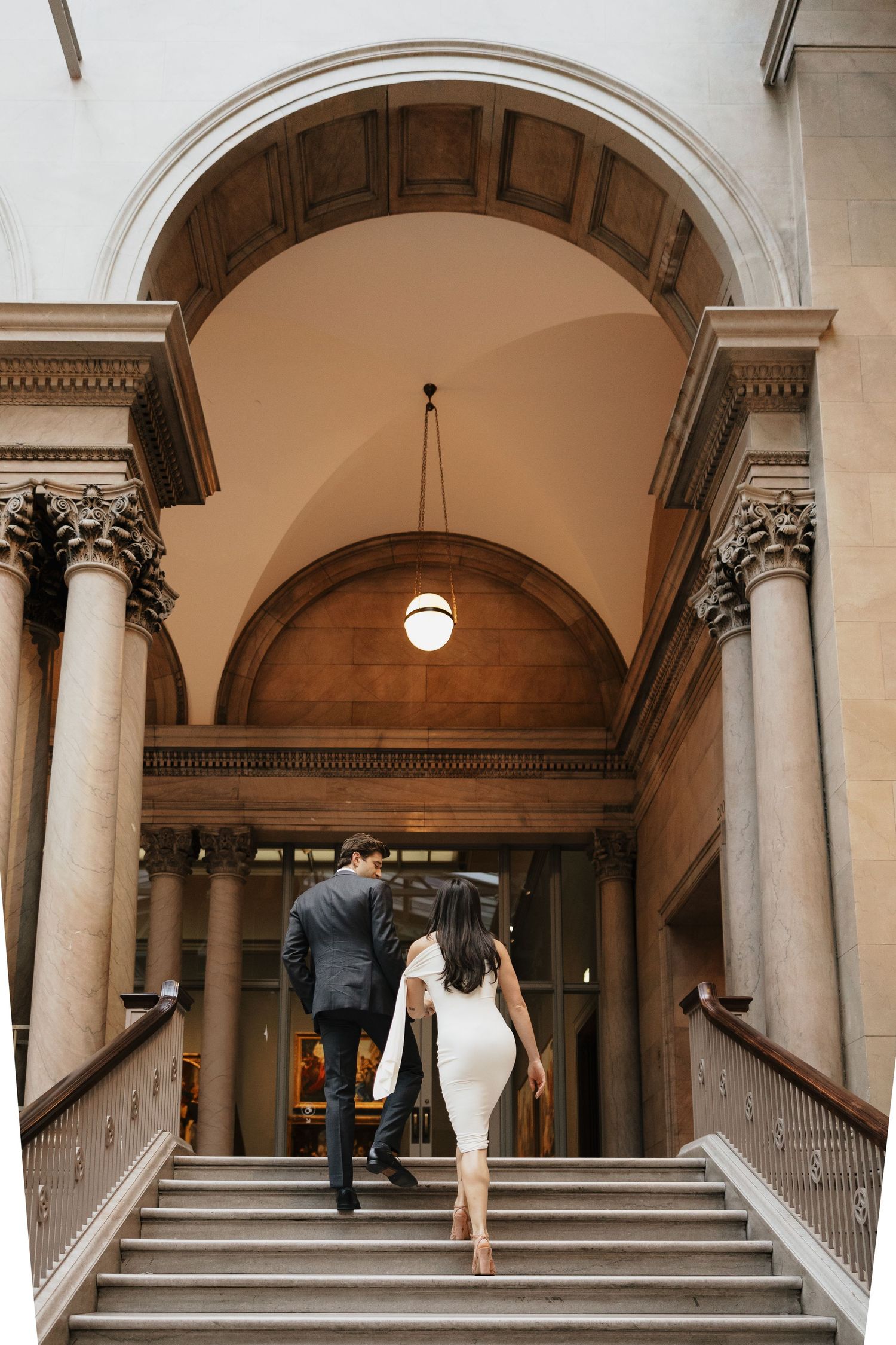 Couple in formal attire walking up a grand marble staircase toward an arched entrance with stone columns and a hanging pendant light.
