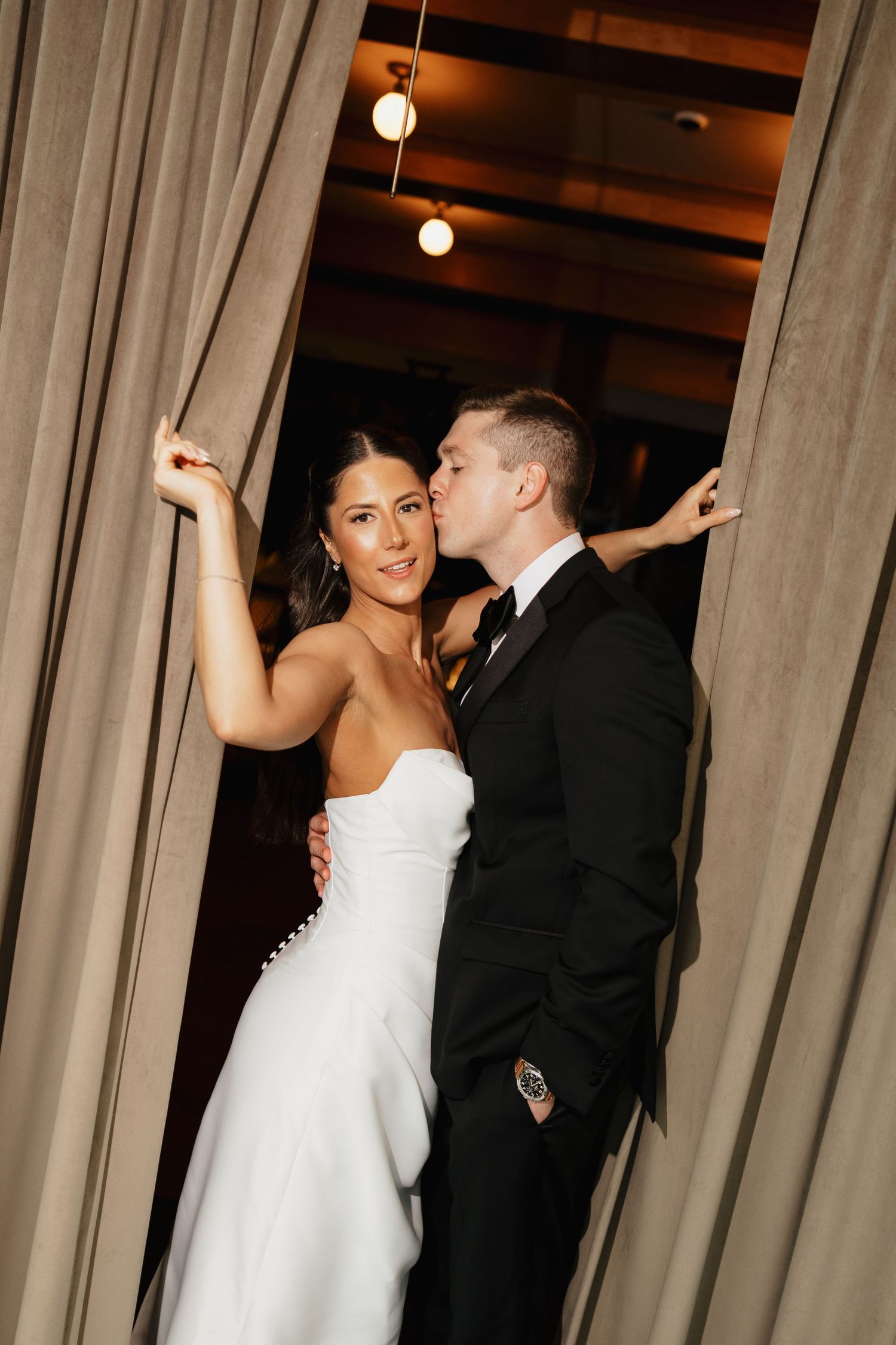 A groom in a black suit kisses the cheek of a smiling bride in a strapless white gown as they pose between tan curtains.