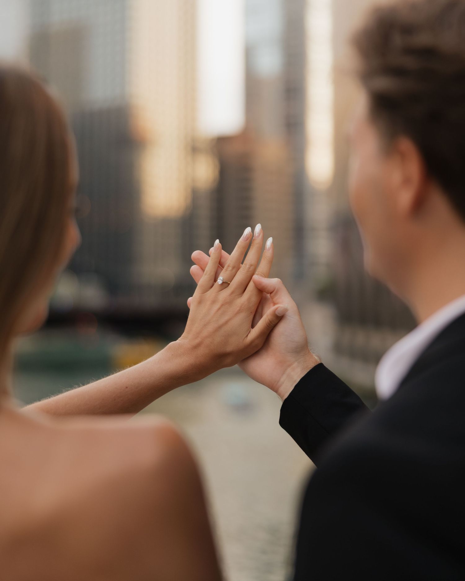 Couple pressing their hands together to show an engagement ring, with a blurred city skyline glowing behind them at sunset.