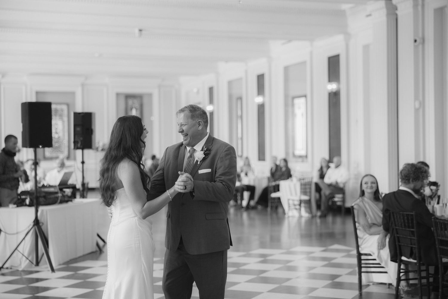 A couple shares their first dance in an elegant white ballroom with tall windows and checkered floors.