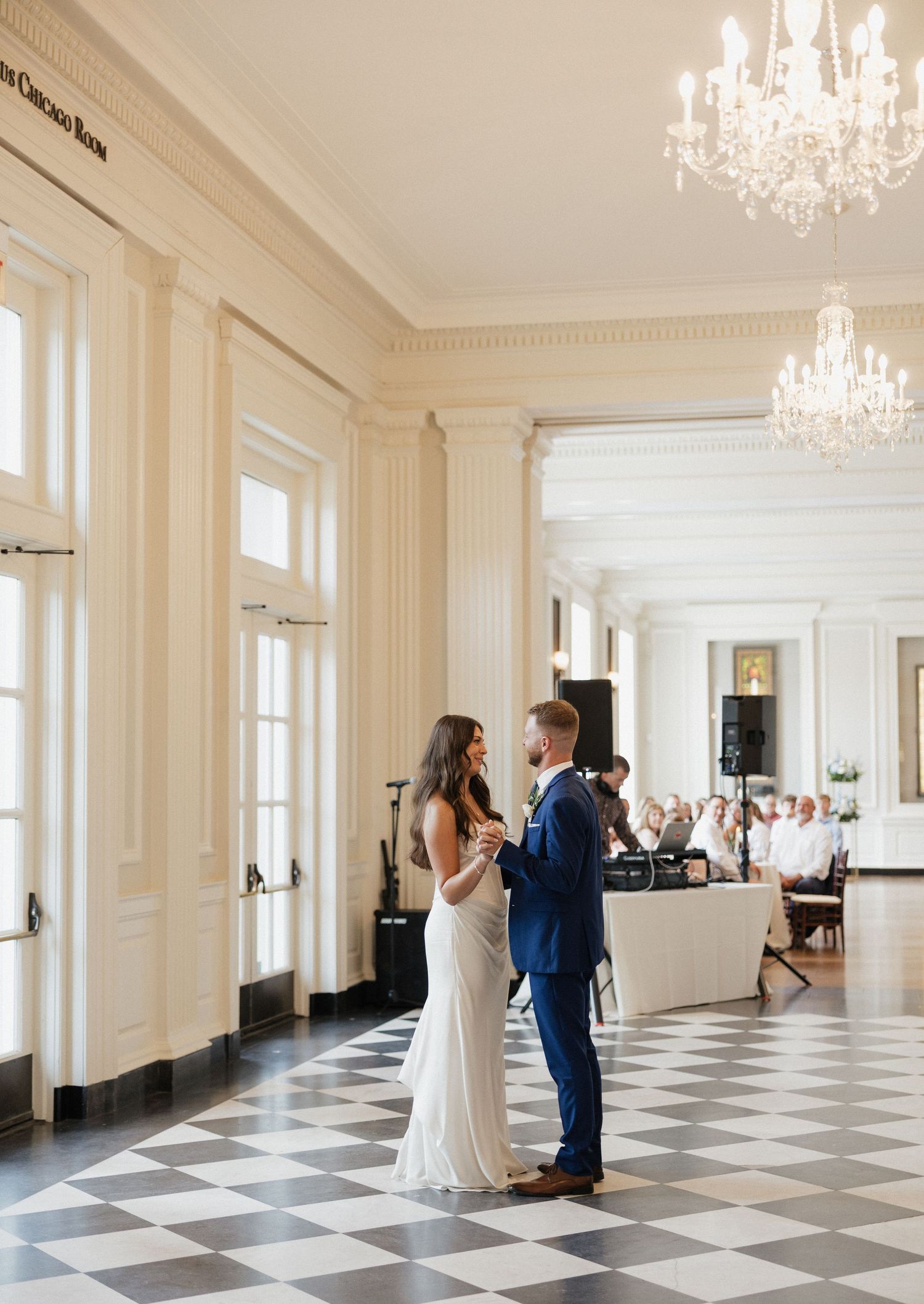 A couple shares their first dance on a black and white checkered floor beneath elegant crystal chandeliers in a grand ballroom.