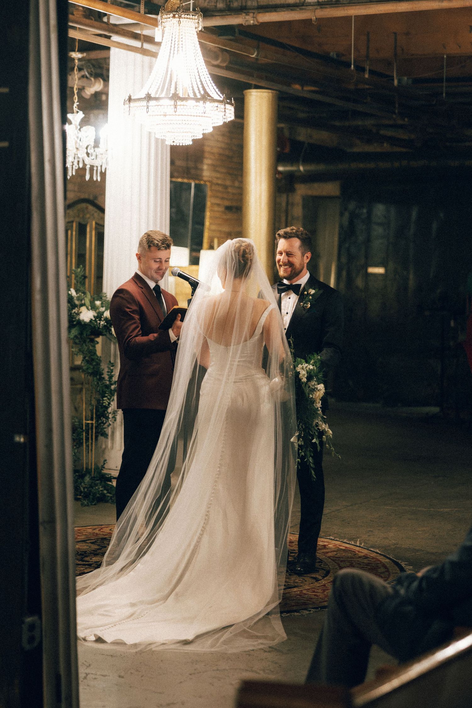A bride in a flowing white dress and veil stands with others in an elegant indoor venue with crystal chandeliers.
