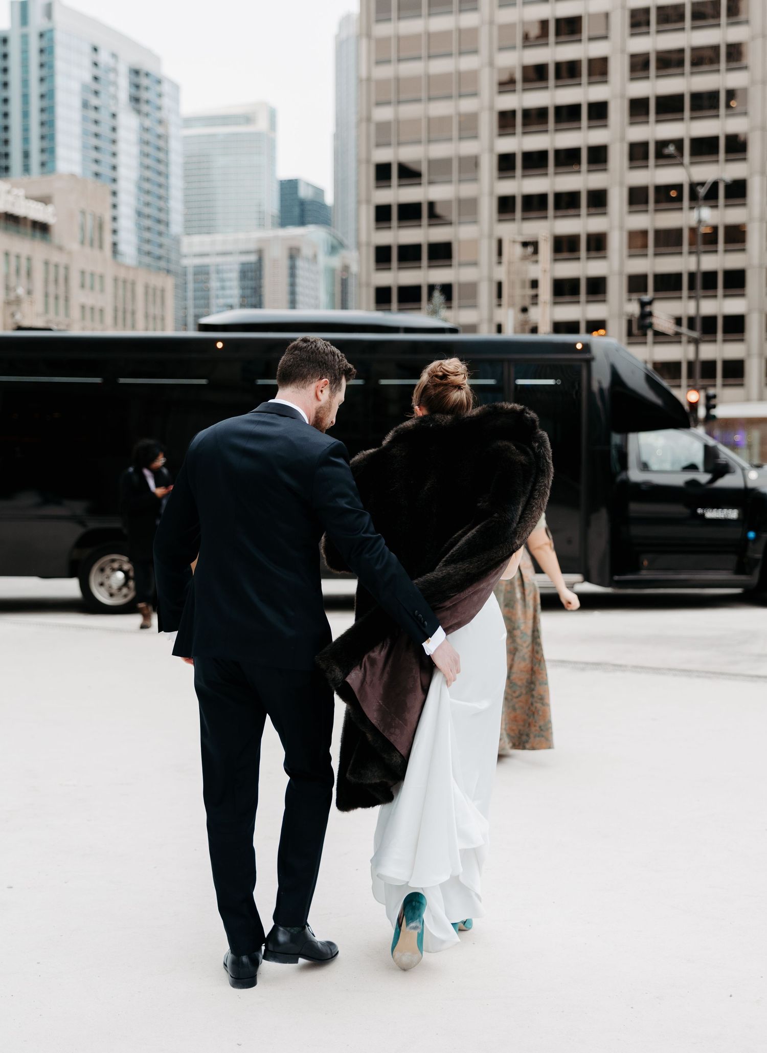 A couple in formal wedding attire walks together on a rooftop with city buildings in the background.
