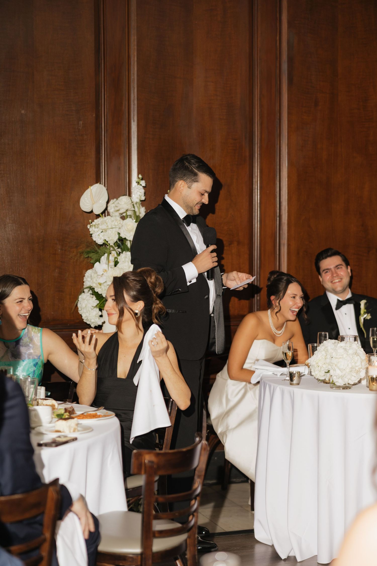 Groom in tuxedo giving wedding speech at elegant reception while bride and guests laugh at candlelit dinner table.
