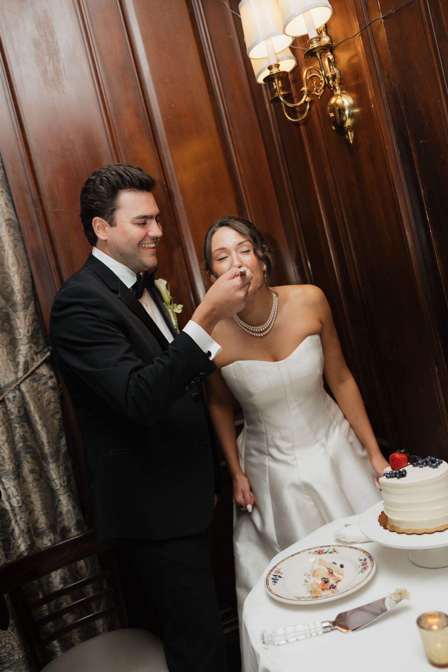 Groom feeding wedding cake to bride in white strapless gown at elegant reception with wood paneling.