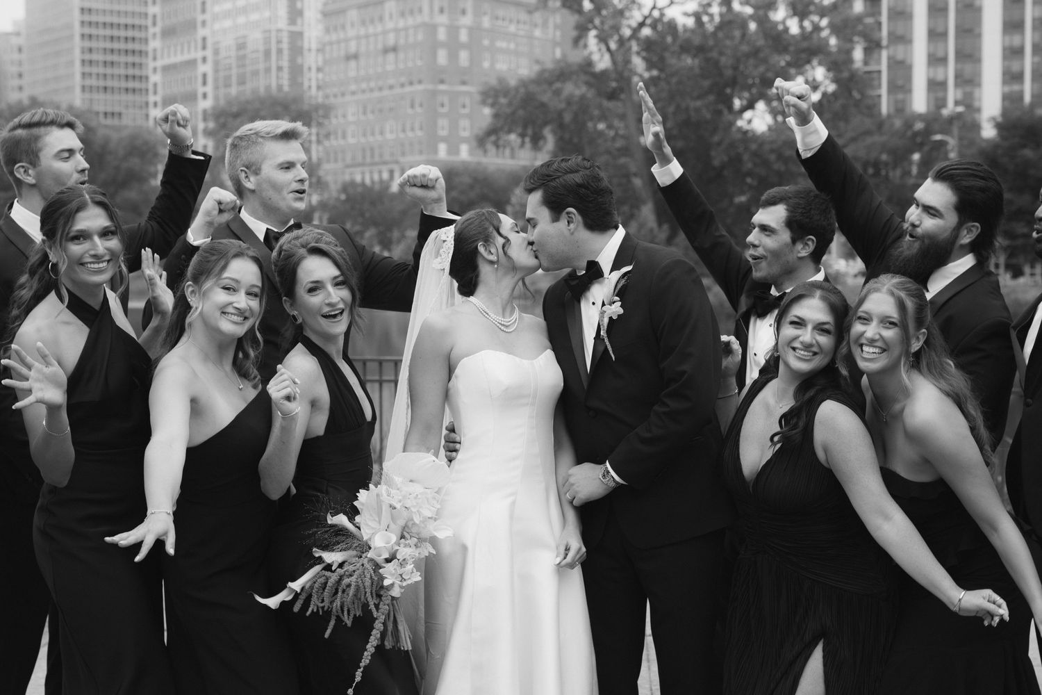 Black and white wedding photo of bride and groom kissing surrounded by cheering wedding party in formal attire outdoors.