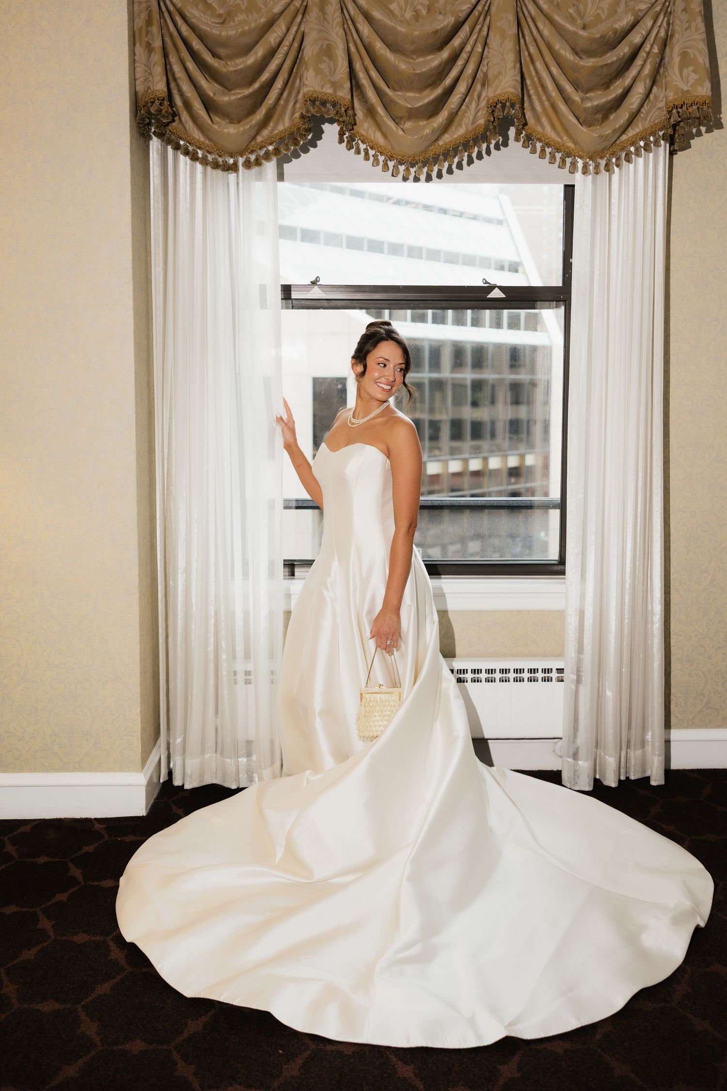 Elegant bride in a strapless white satin gown with a long train poses by a window in a luxurious hotel room.