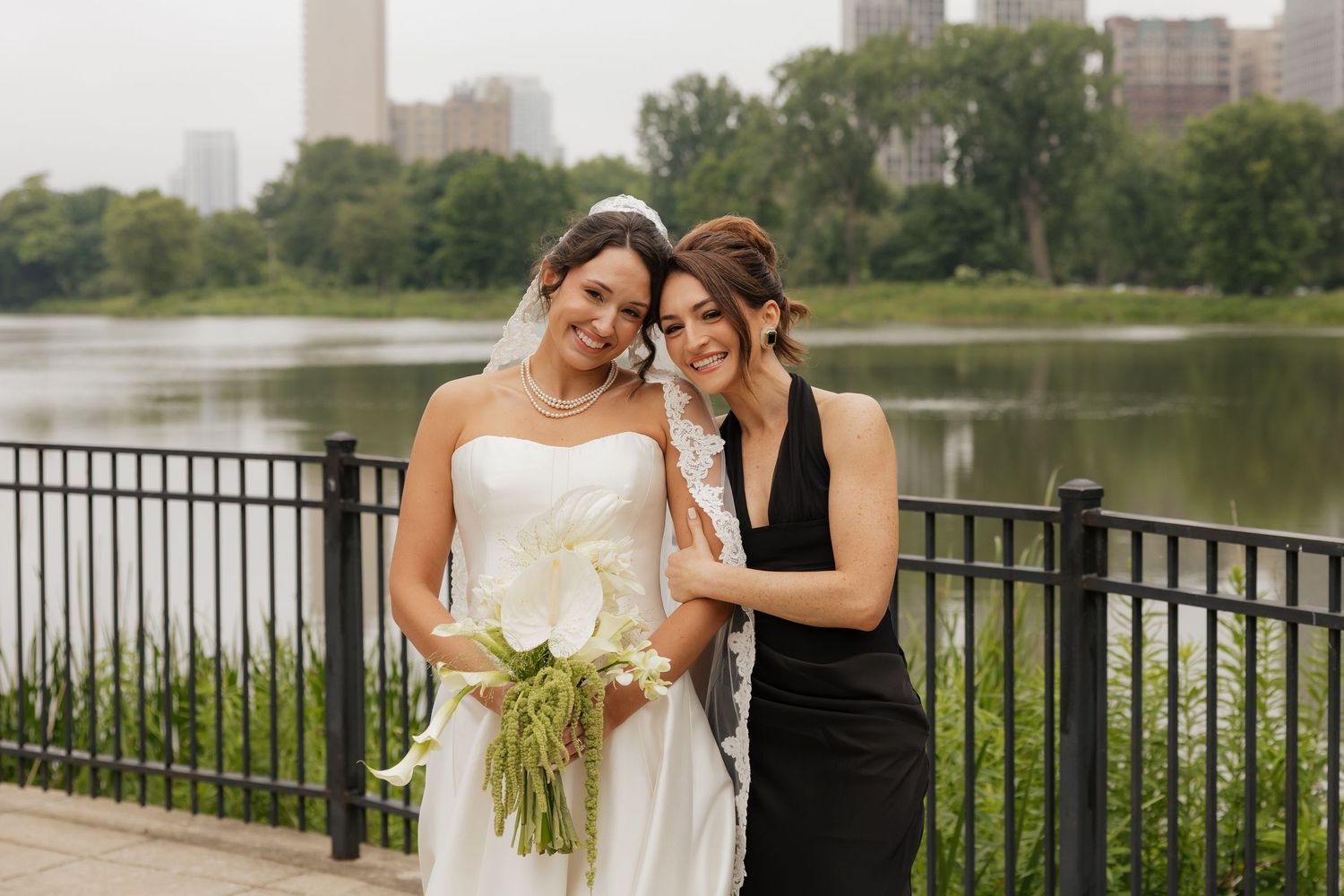Bride in white gown holding bouquet poses with woman in black dress by lakeside railing with city skyline backdrop.