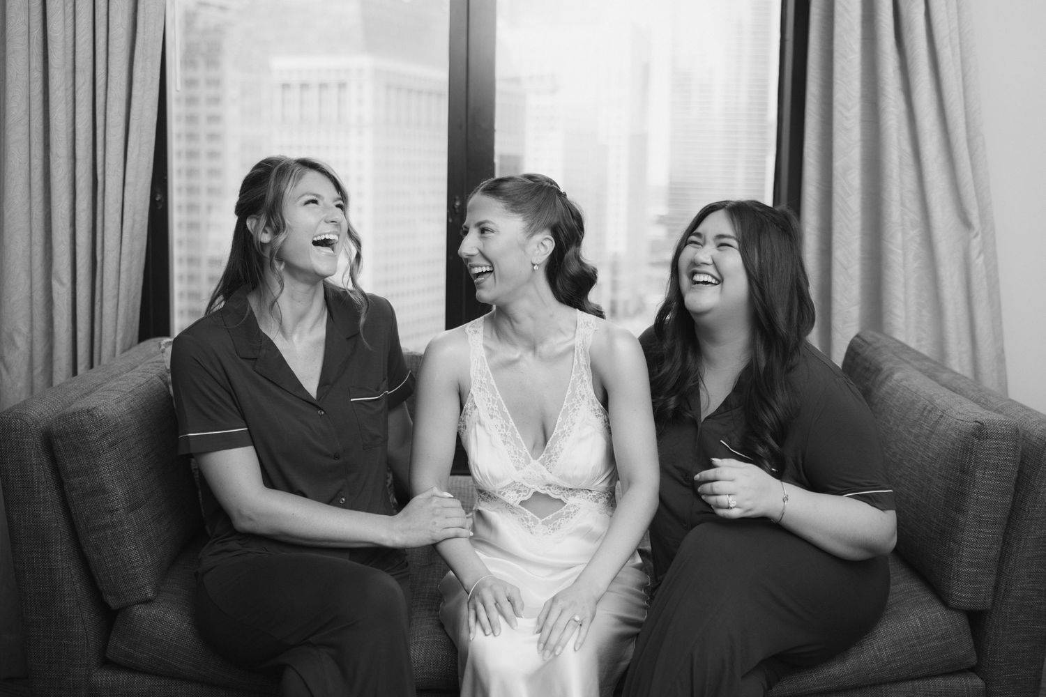 Black and white photo of a bride in white dress laughing with two bridesmaids on a couch near a window.