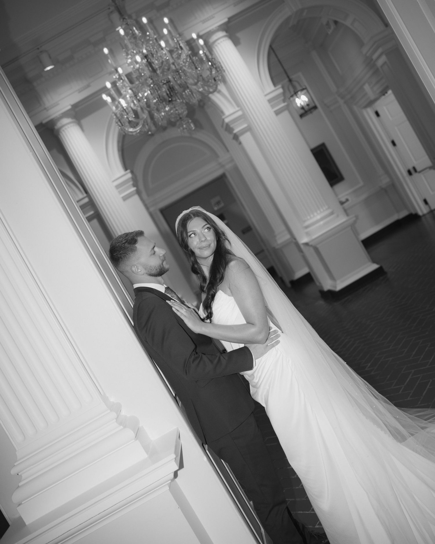A couple shares a romantic dance moment in an elegant ballroom with crystal chandeliers and classic white columns.