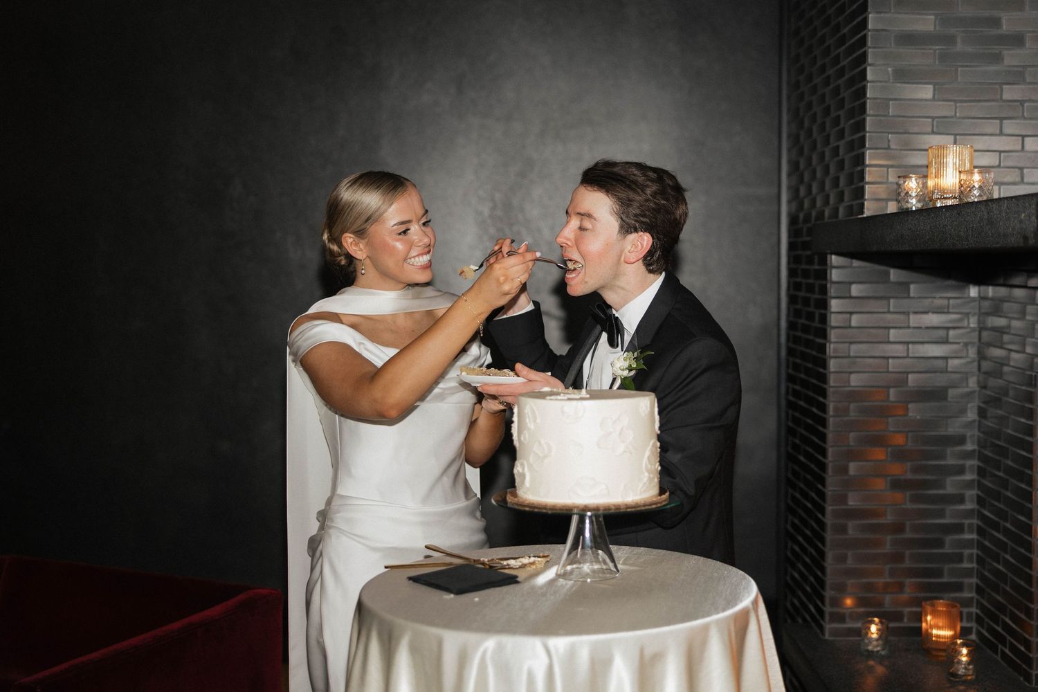 Bride in white dress feeds wedding cake to groom in tuxedo at elegant reception table with candles.