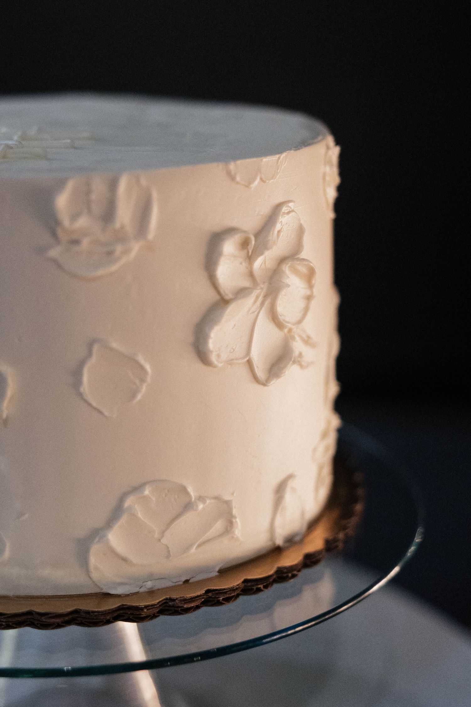 Elegant white frosted cake with textured floral designs on a glass cake stand against dark background.