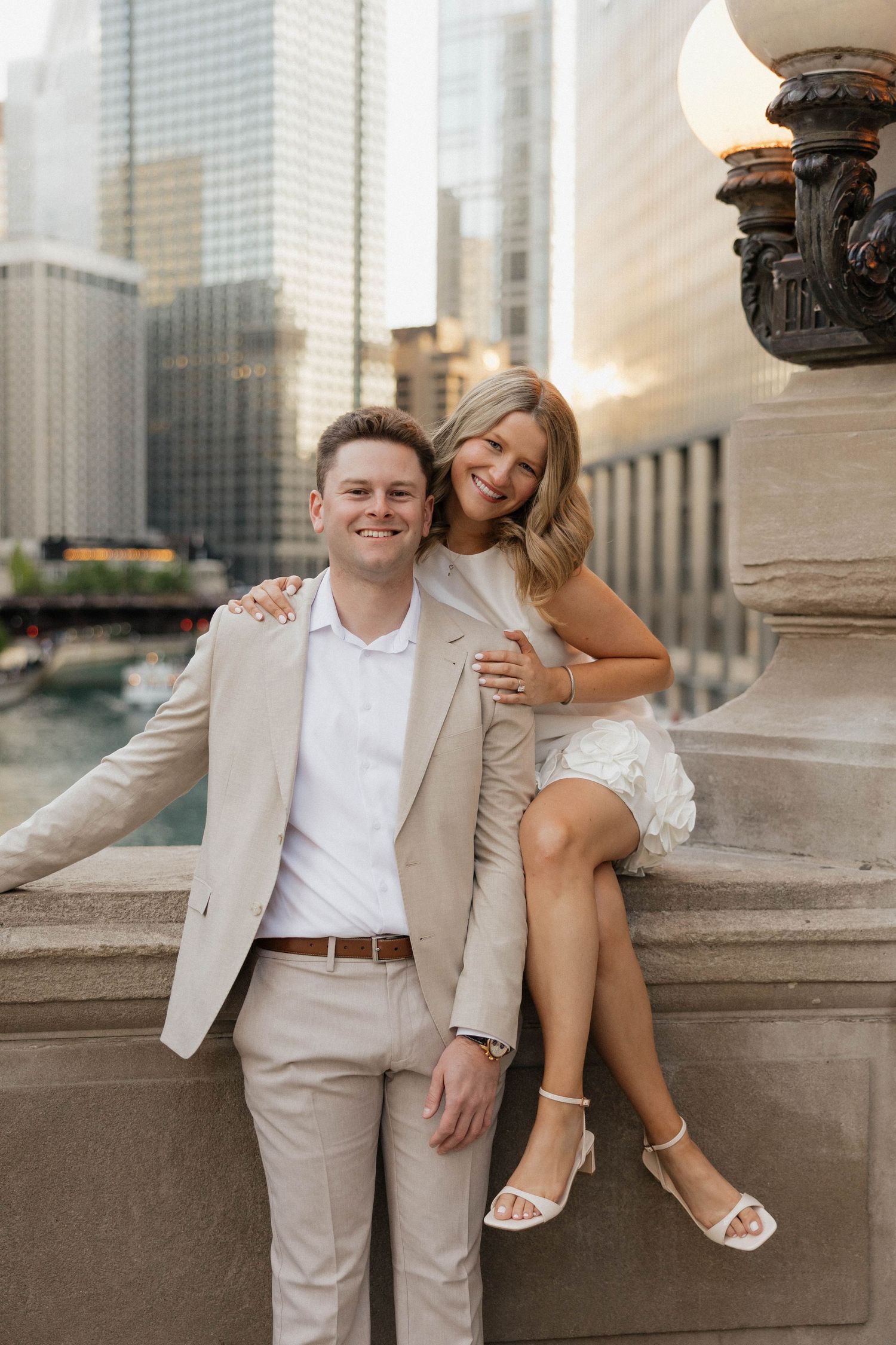 Happy couple posing by Chicago River, man in tan suit and woman in white mini dress smiling, golden hour city skyline behind them.