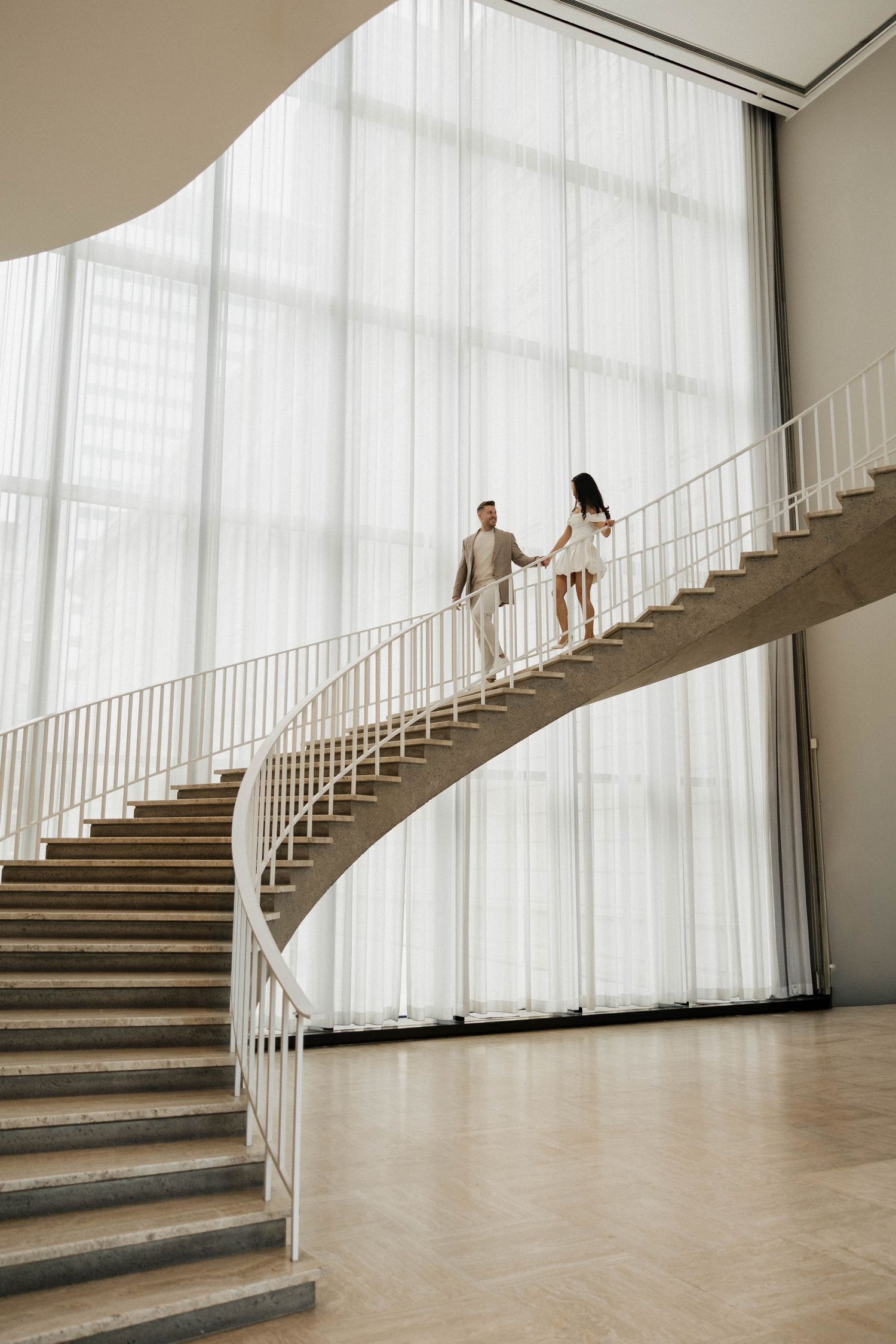 Couple holding hands walking up a curved spiral staircase in a modern building with floor-to-ceiling windows.