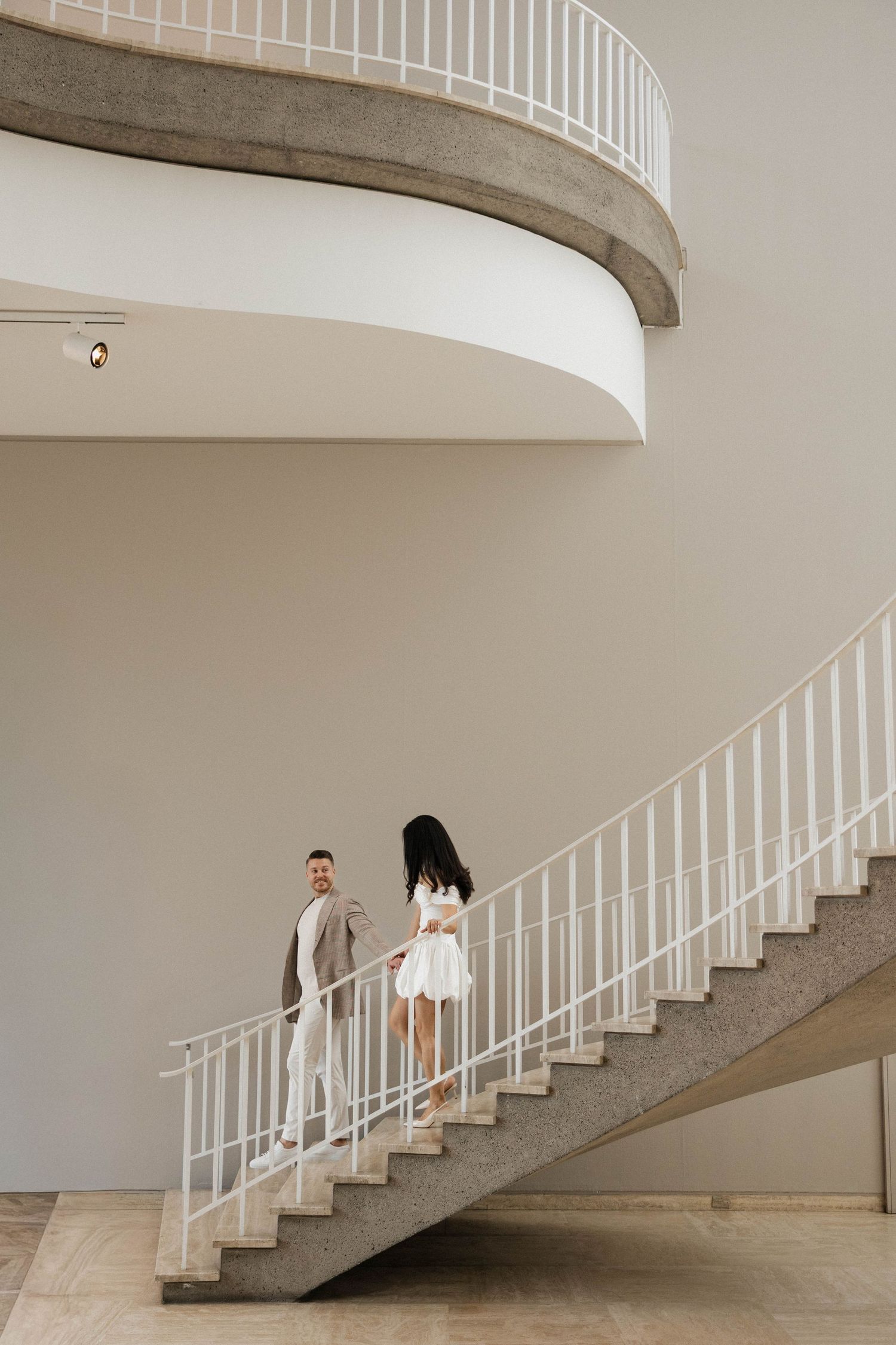 Man and woman in white outfits descending a curved concrete staircase in a minimalist modern interior space.