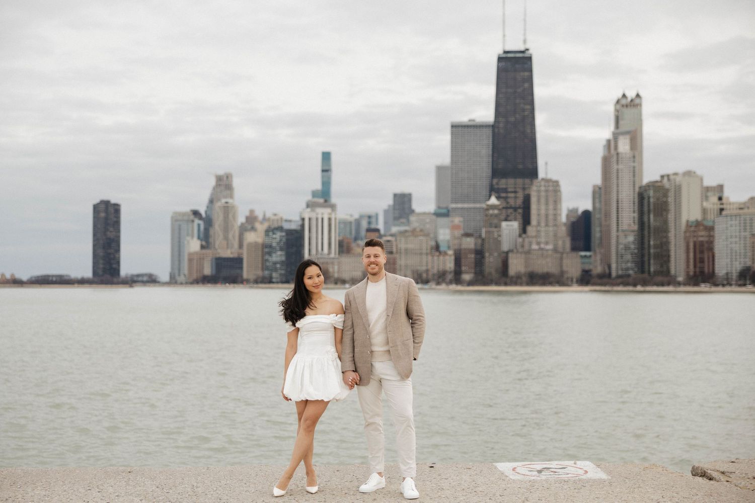 Couple poses by Lake Michigan with Chicago skyline and John Hancock Center in background on cloudy day.