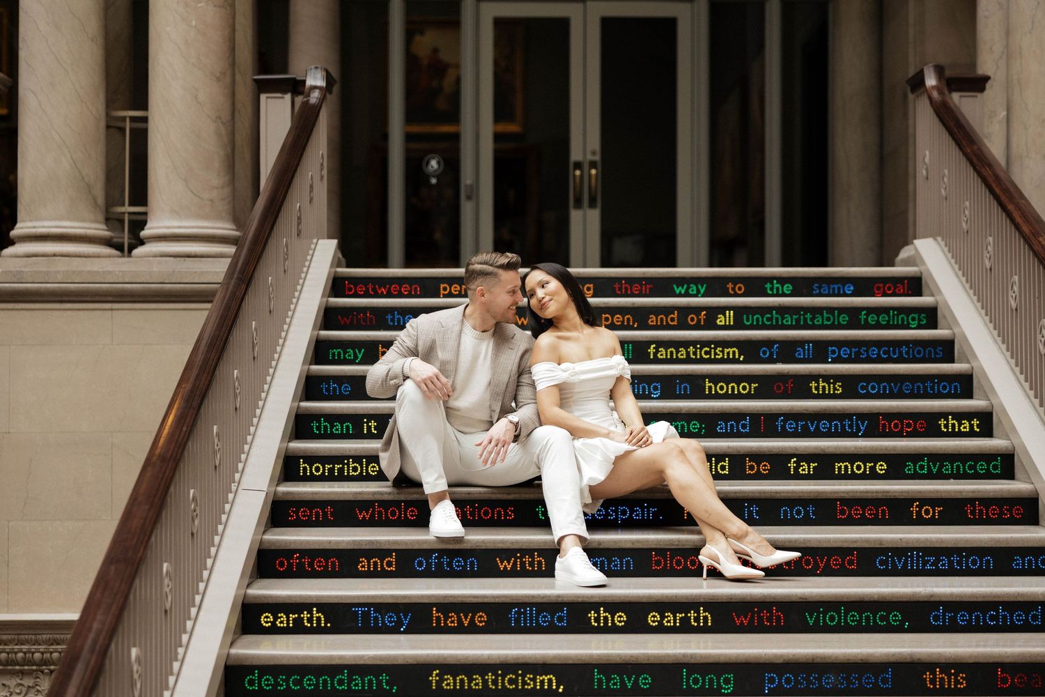 Couple sitting together on colorful text-covered steps of a grand building, woman in white dress, man in light suit.