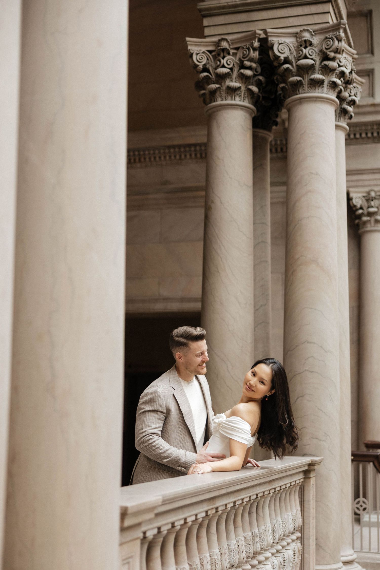 Elegant couple posing on a classical stone balcony with Corinthian columns during a romantic engagement photo session.