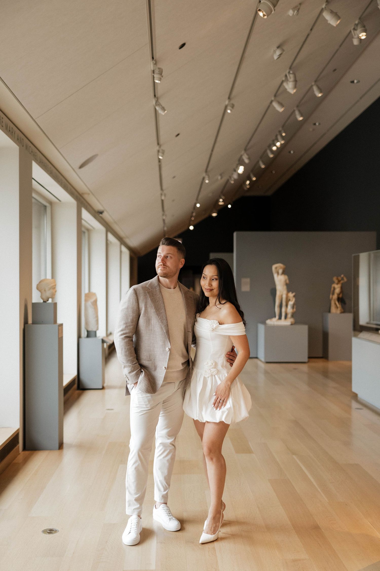 Couple posing in a museum gallery, man in grey blazer and woman in white off-shoulder mini dress.