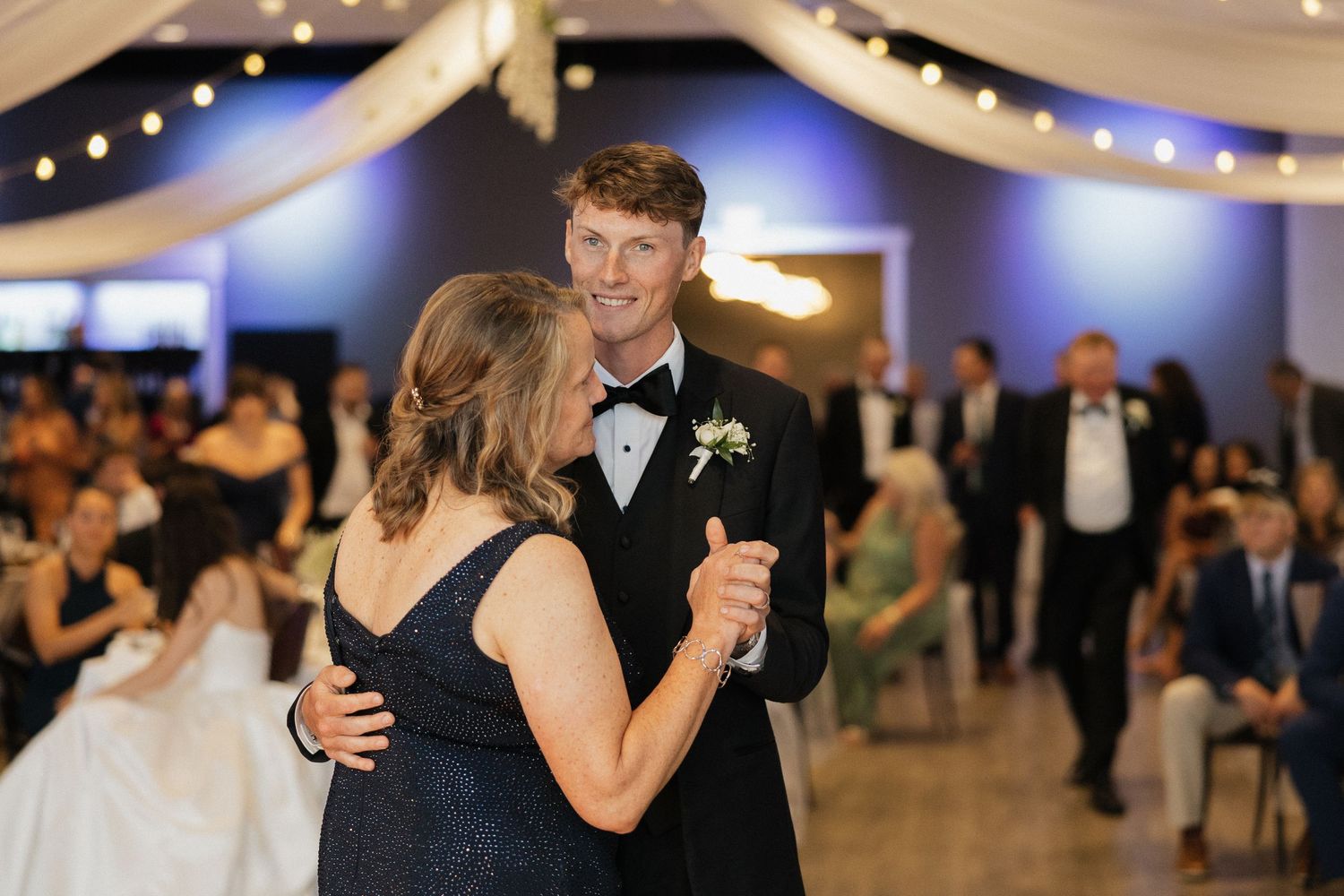 Groom in black tuxedo shares mother-son dance at elegant wedding reception with blue lighting and string lights.