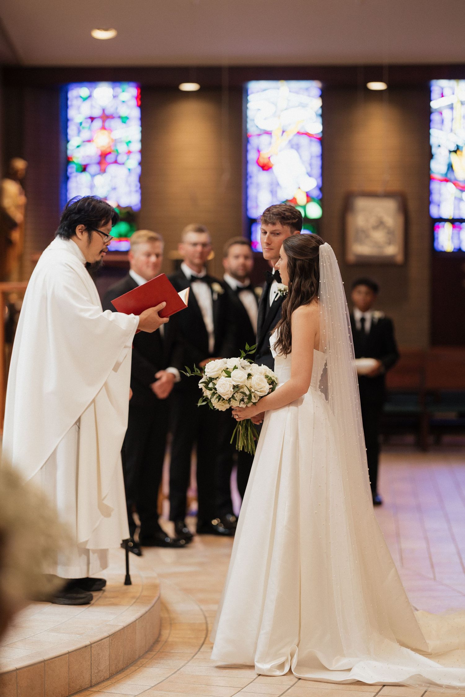 Bride and groom exchange vows before a priest in a church with colorful stained glass windows during wedding ceremony.