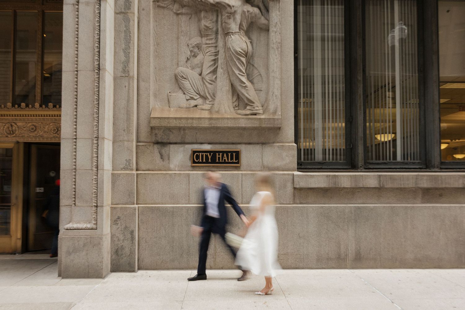 Two blurred figures walk past a grand stone City Hall building with ornate sculptures above the entrance.