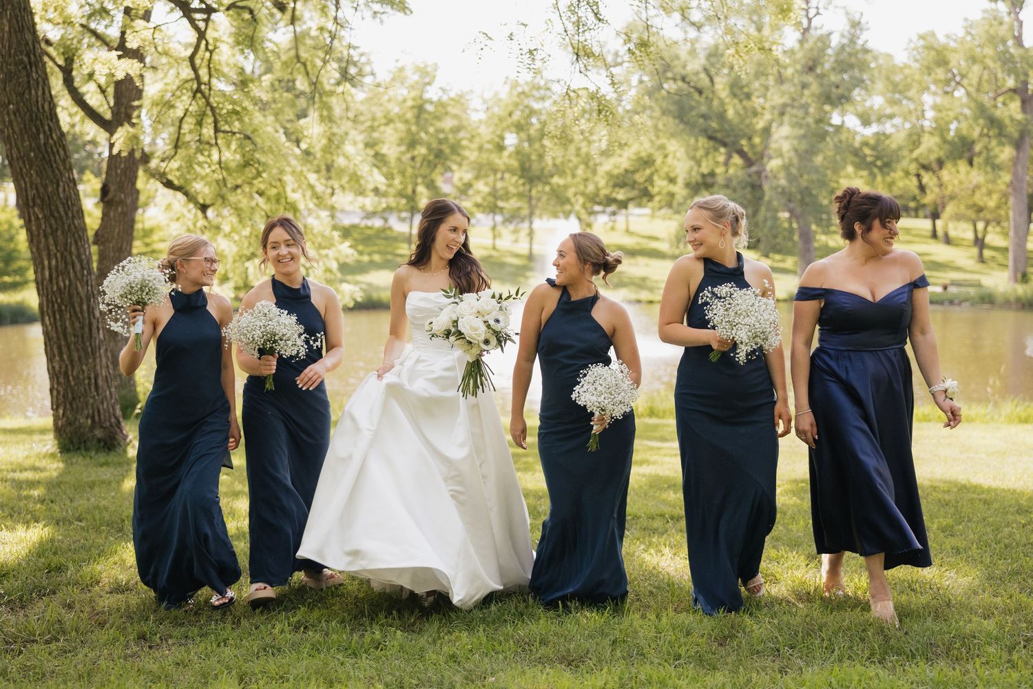 Bride in white gown walks with five bridesmaids in navy blue dresses holding white bouquets outdoors.