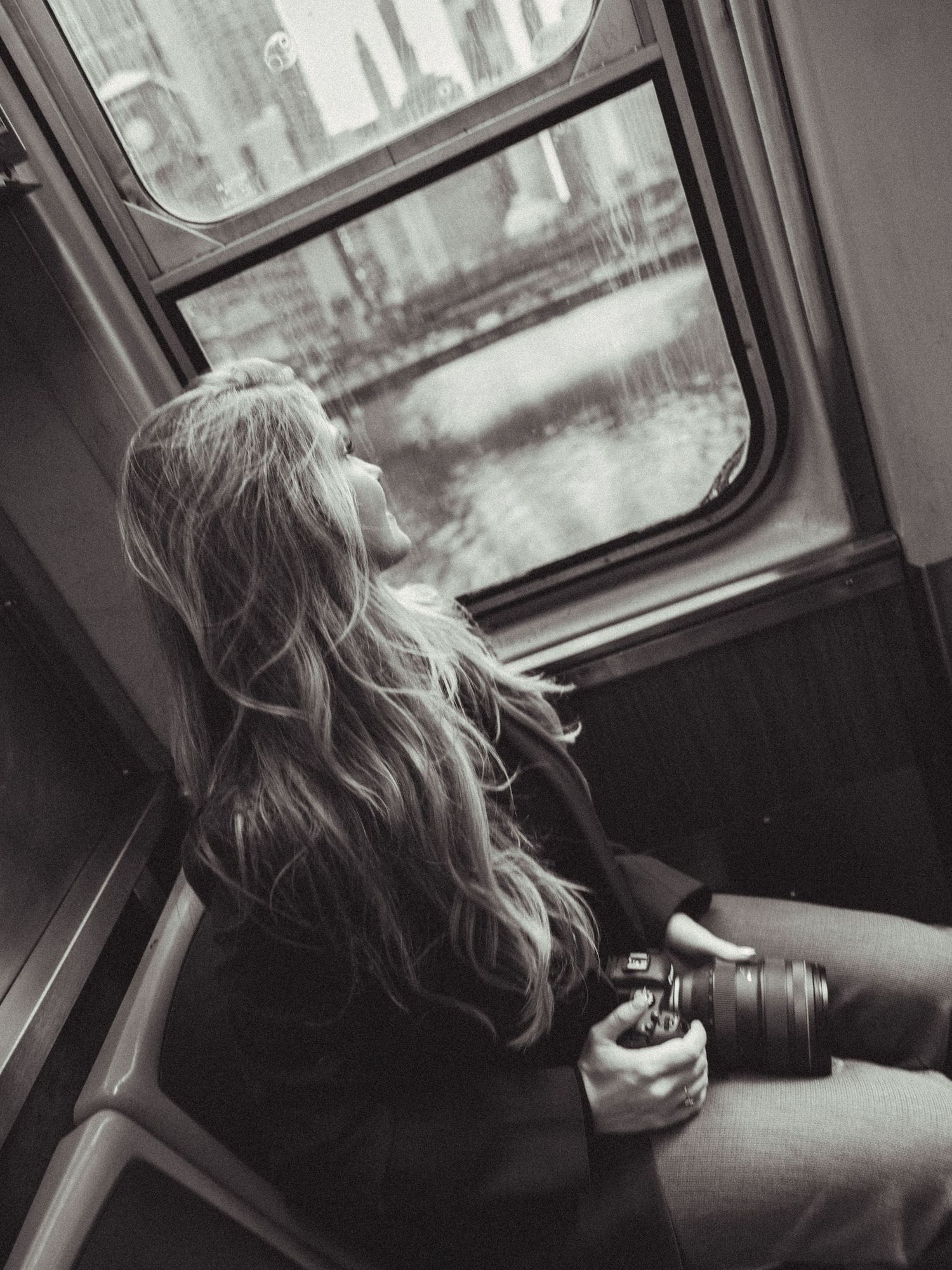 Black and white photo of a woman with long hair holding a camera while gazing out a train window.