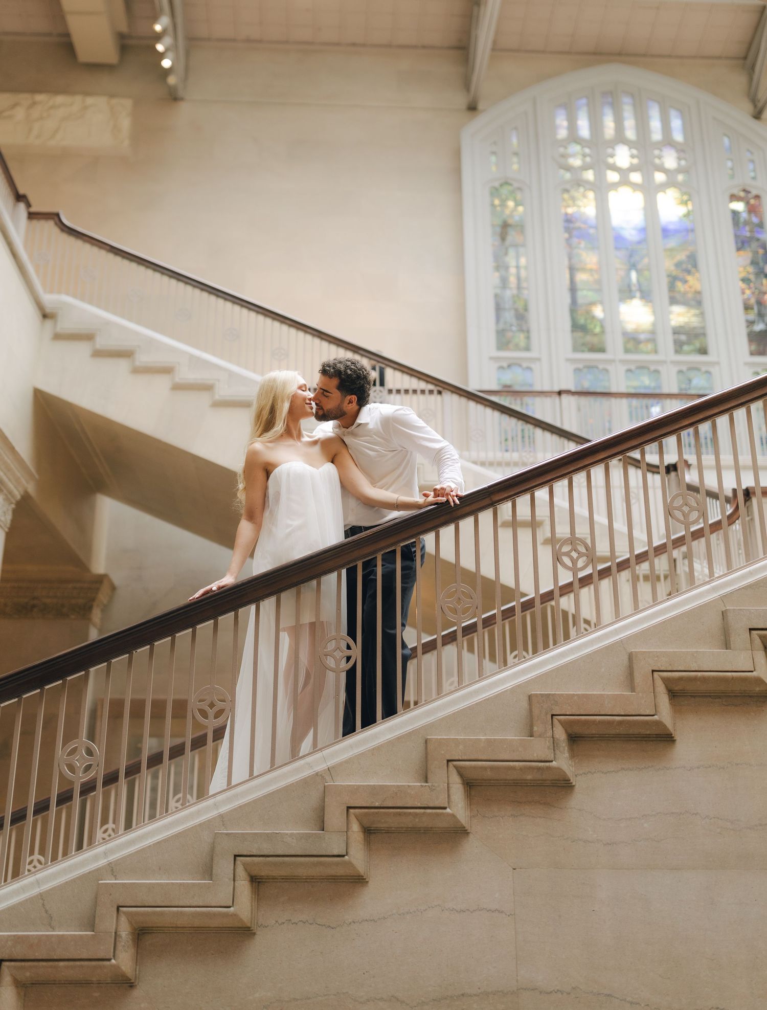 Couple sharing a romantic kiss on an elegant staircase with ornate railings and stained glass window backdrop.