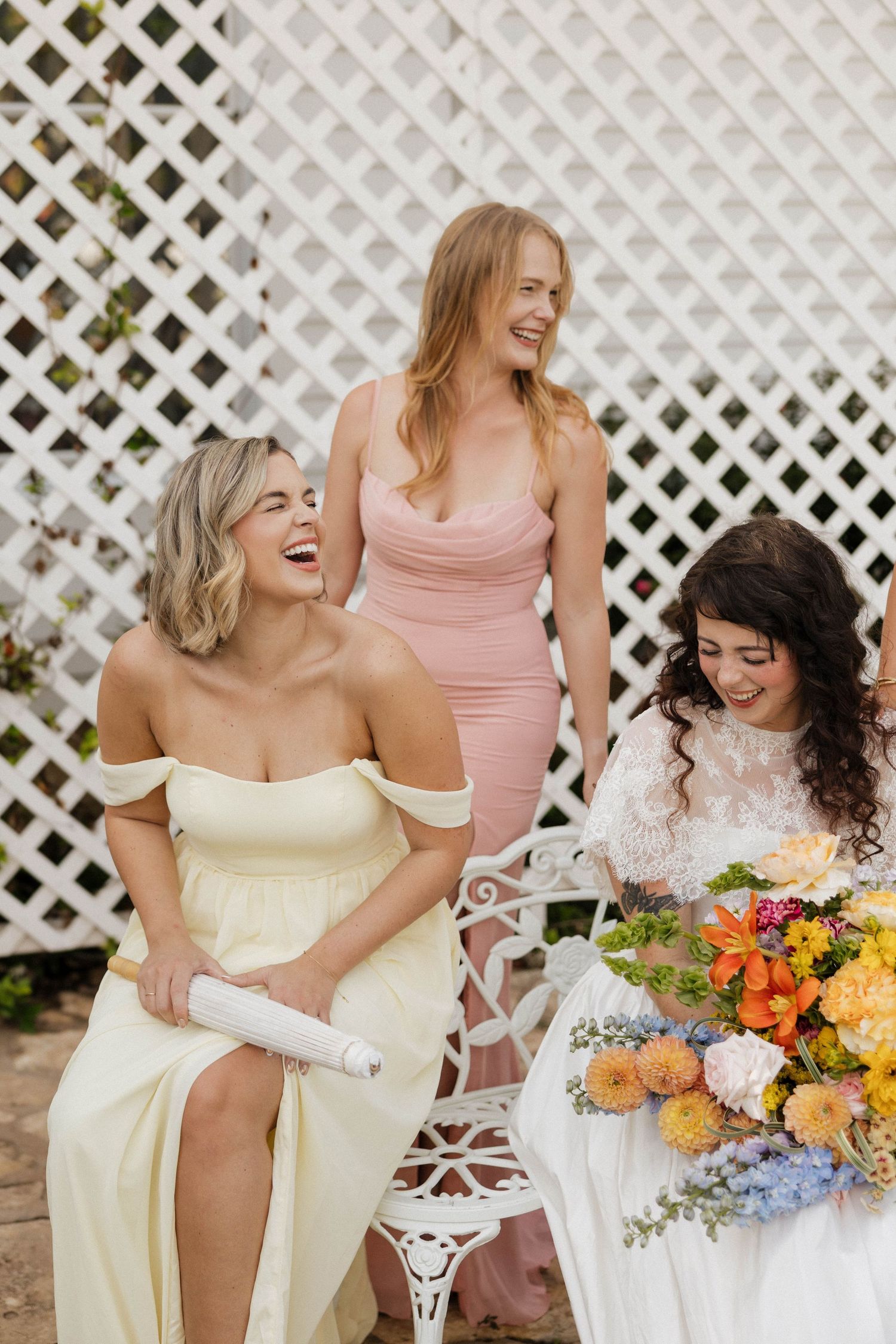 Three women in elegant dresses laugh together at an outdoor wedding, surrounded by colorful floral arrangements.