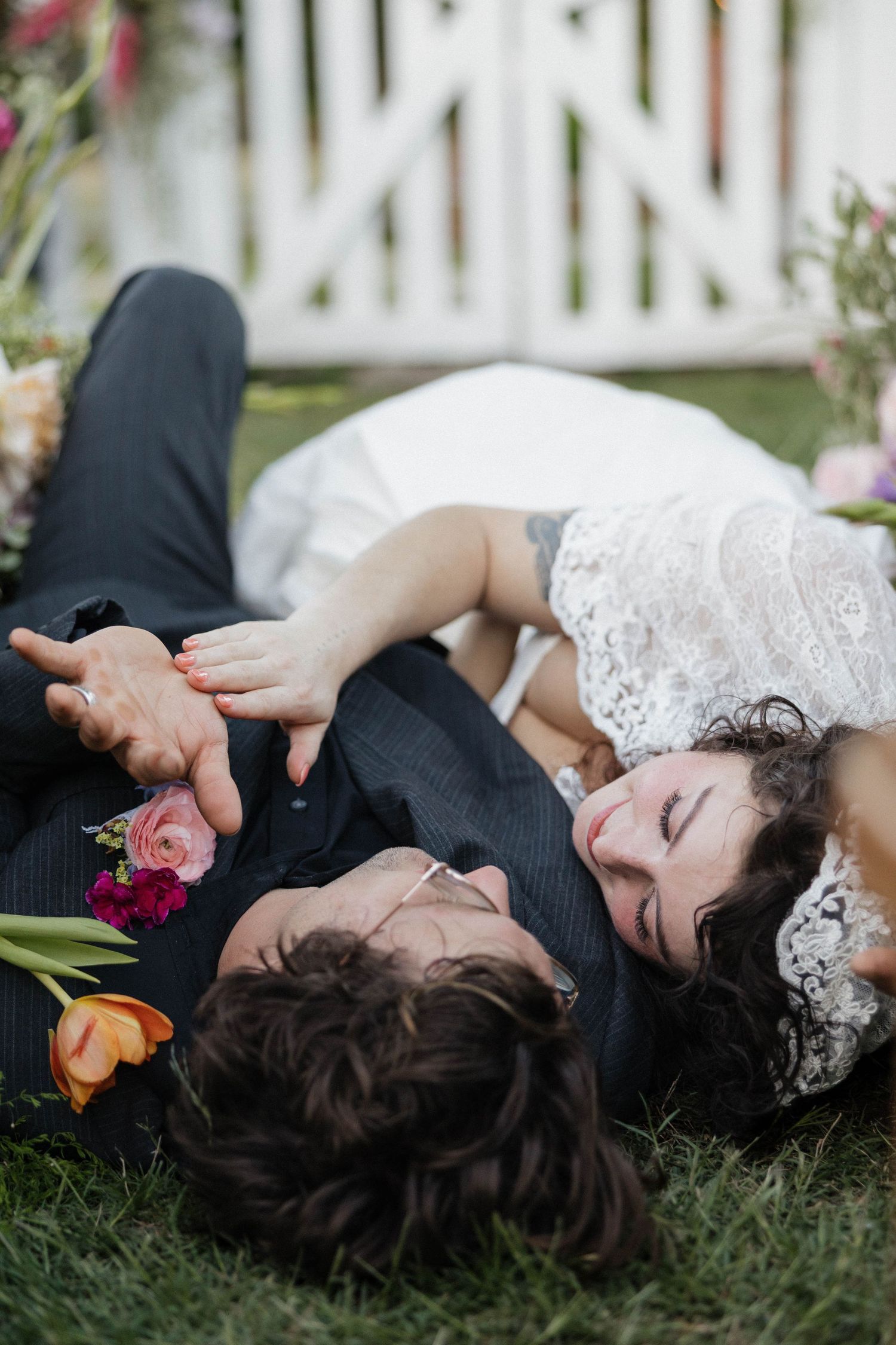Bride and groom lying on grass surrounded by colorful flowers, sharing a romantic moment on their wedding day.