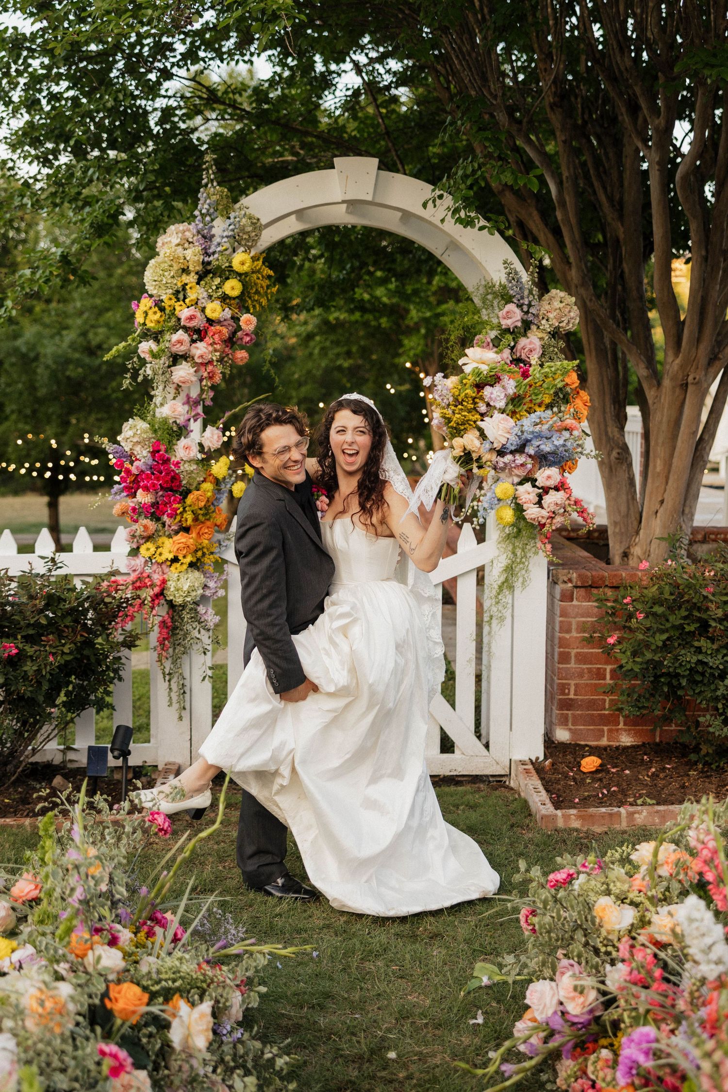 Joyful bride and groom laughing under a colorful floral arch with vibrant blooms at an outdoor garden wedding ceremony.