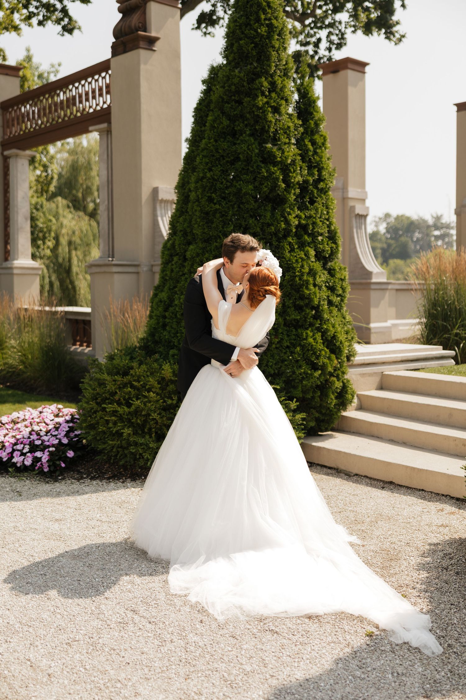 Bride and groom share a romantic kiss in a beautiful garden setting, with the bride wearing a flowing white ball gown.