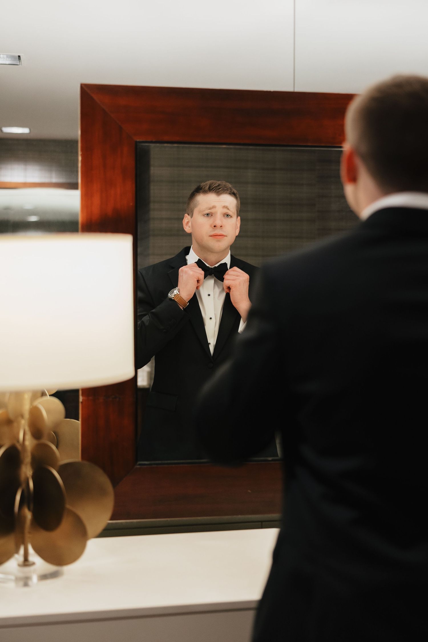 Groom in black tuxedo adjusting bow tie while looking at his reflection in a wooden-framed mirror.