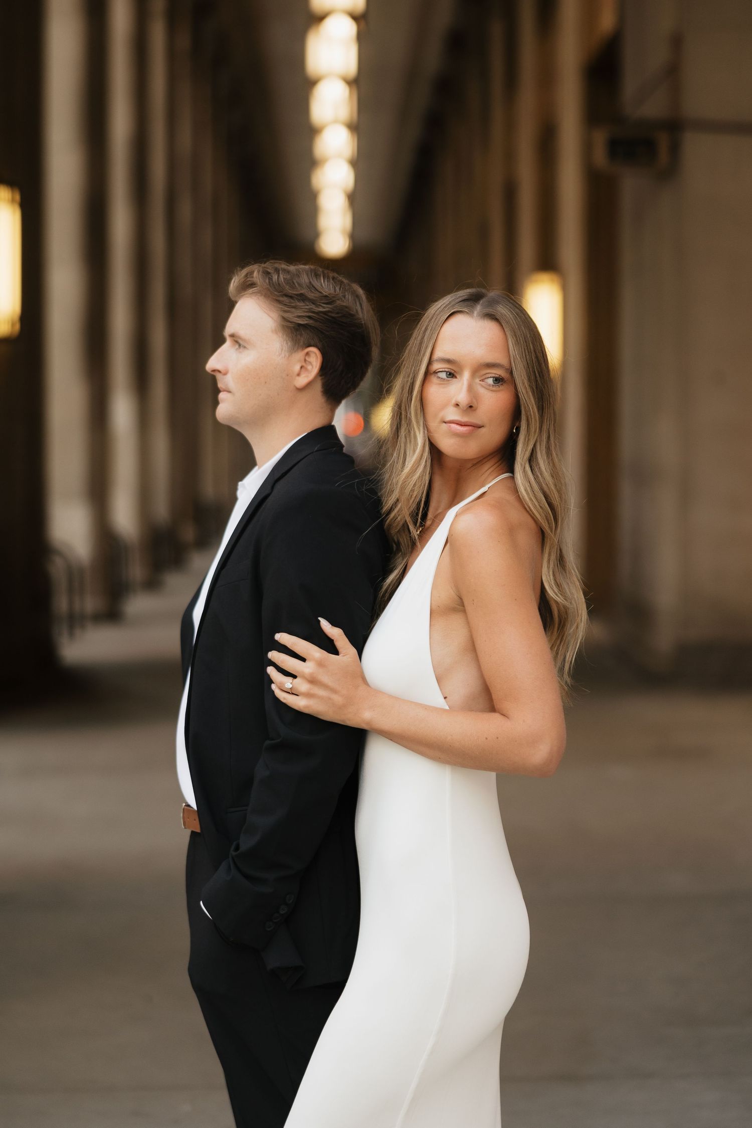 Couple in elegant attire, woman in white gown and man in black suit, posing in a grand architectural hallway.