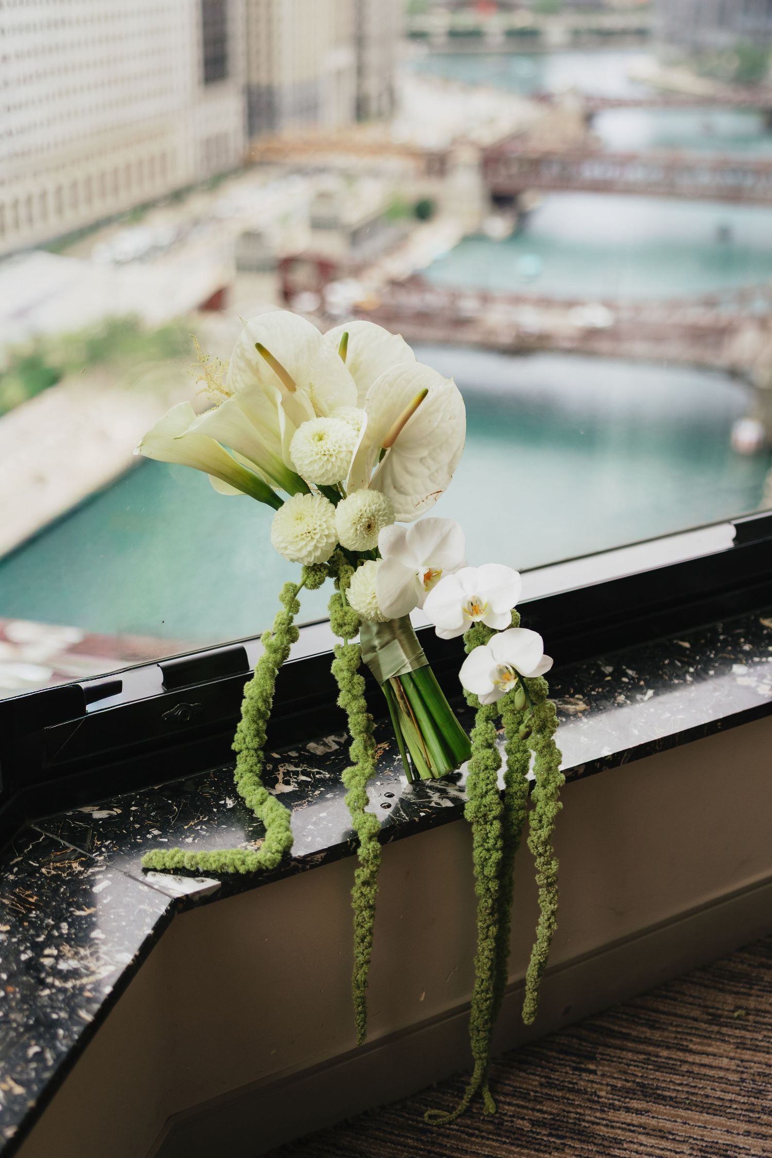 Elegant white bridal bouquet with calla lilies, orchids, and trailing green amaranthus resting on a windowsill overlooking a city river.