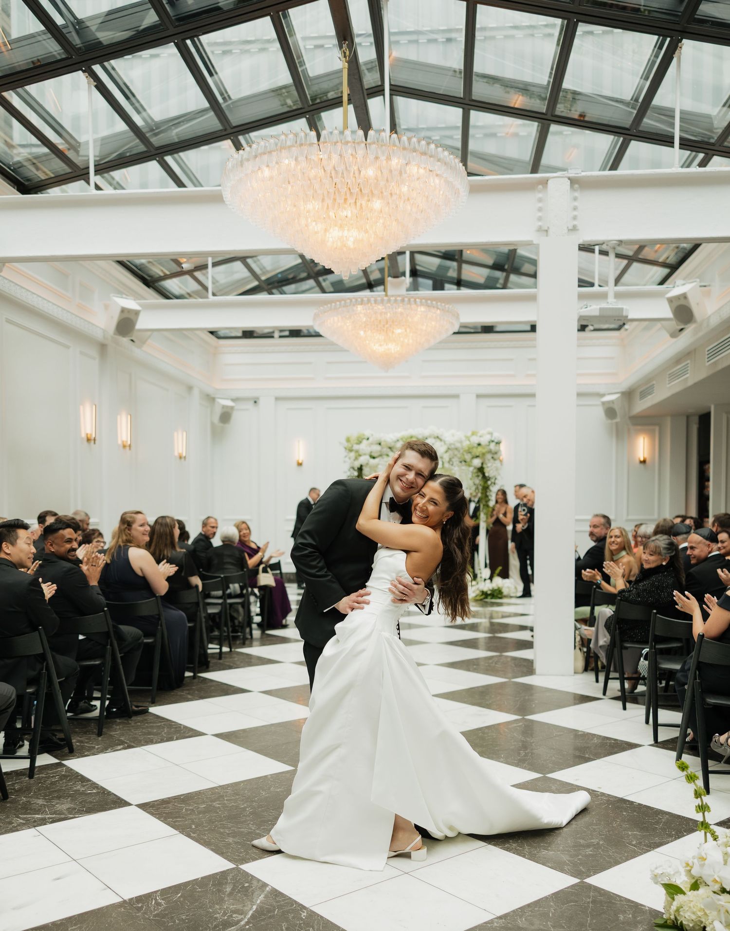 Bride and groom share a romantic dip kiss on a black and white checkered dance floor beneath elegant chandeliers.