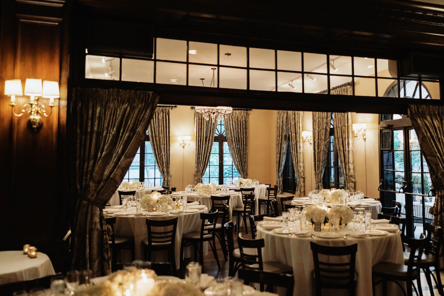 Elegant wedding reception venue with round tables, white linens, dark curtains, and warm lighting in a classic ballroom setting.