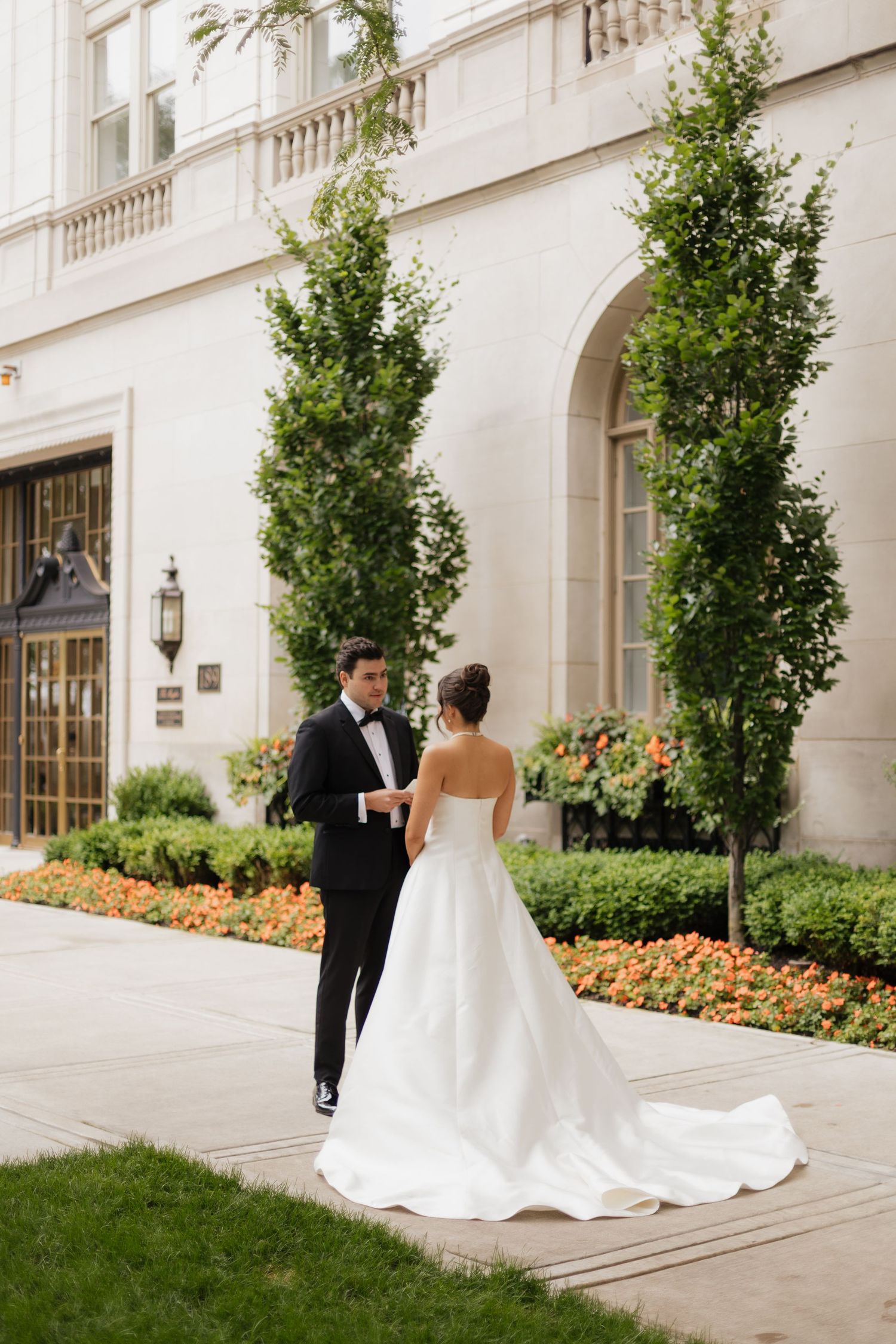 A bride in a white gown and groom in a black tuxedo walk hand in hand outside an elegant building with lush greenery.