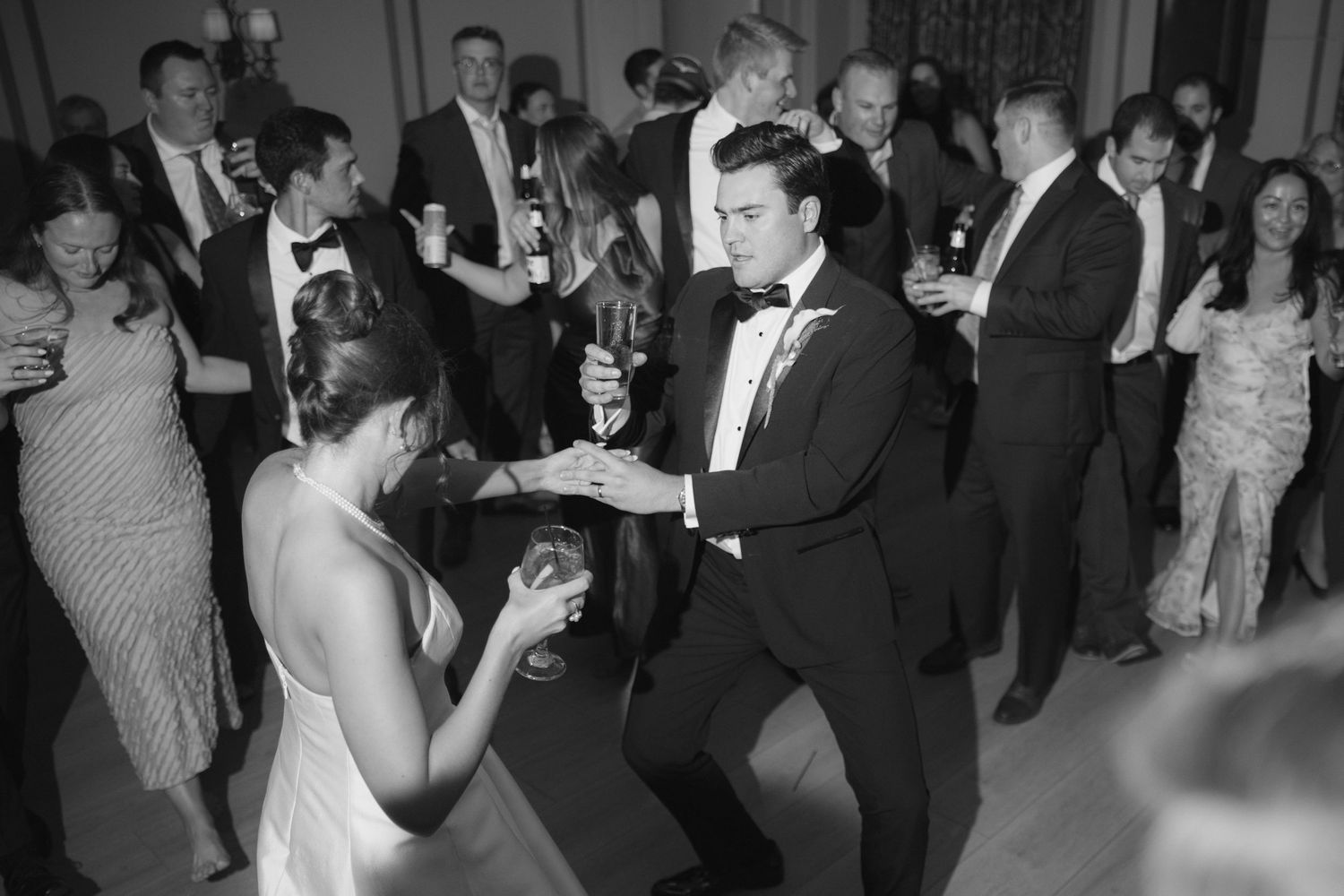 Black and white photo of bride and groom dancing joyfully at their wedding reception surrounded by guests.