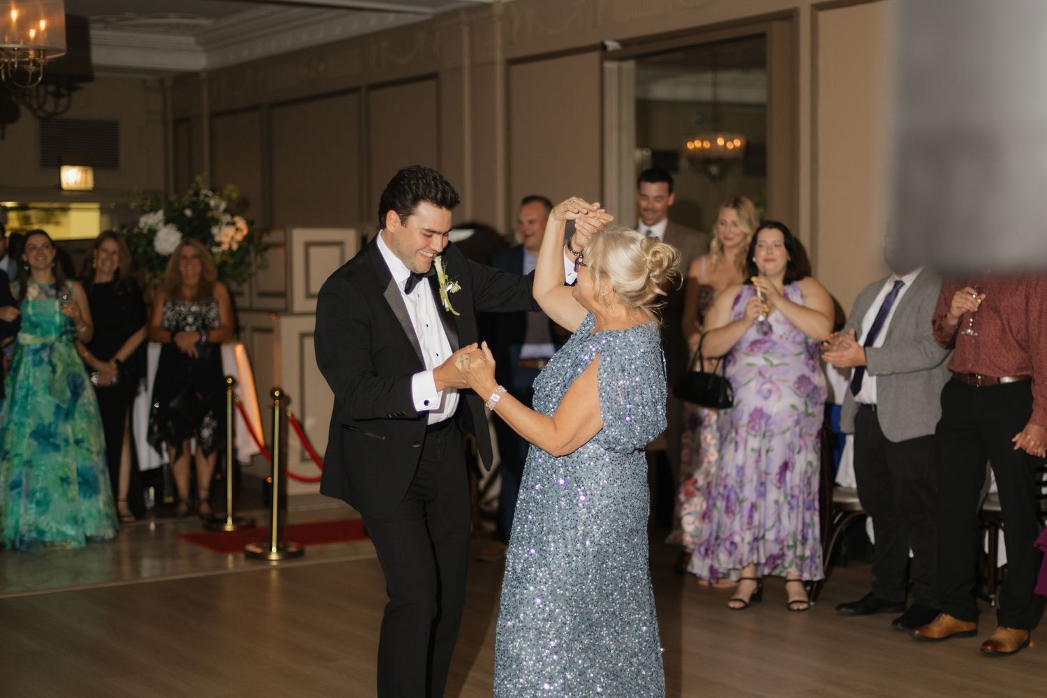 Groom dancing with mother in silver sequin dress at elegant wedding reception with guests watching.